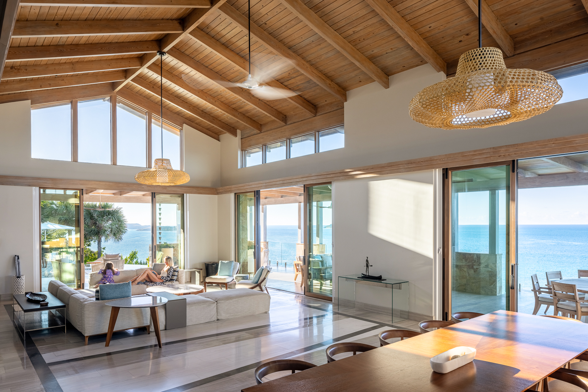 A spacious, modern living room in Maronti Villa boasts high wooden ceilings, large windows, and sliding glass doors showcasing a stunning ocean view. The room features contemporary furniture, including two sofas, a coffee table, a wooden dining table, and woven pendant lights.