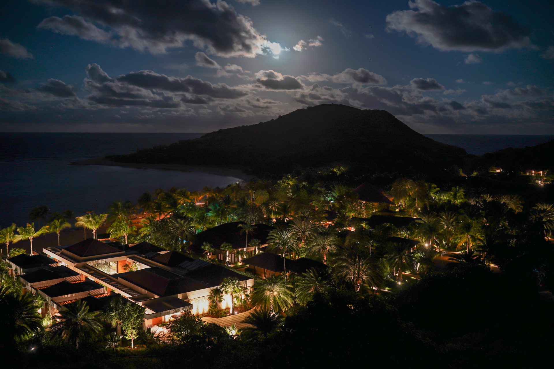 A moonlit tropical resort scene featuring illuminated villas nestled among dense palm trees. The calm ocean water is visible in the background, with a prominent hill silhouetted against a partly cloudy night sky. The tranquil night ambience highlights the serene landscape.