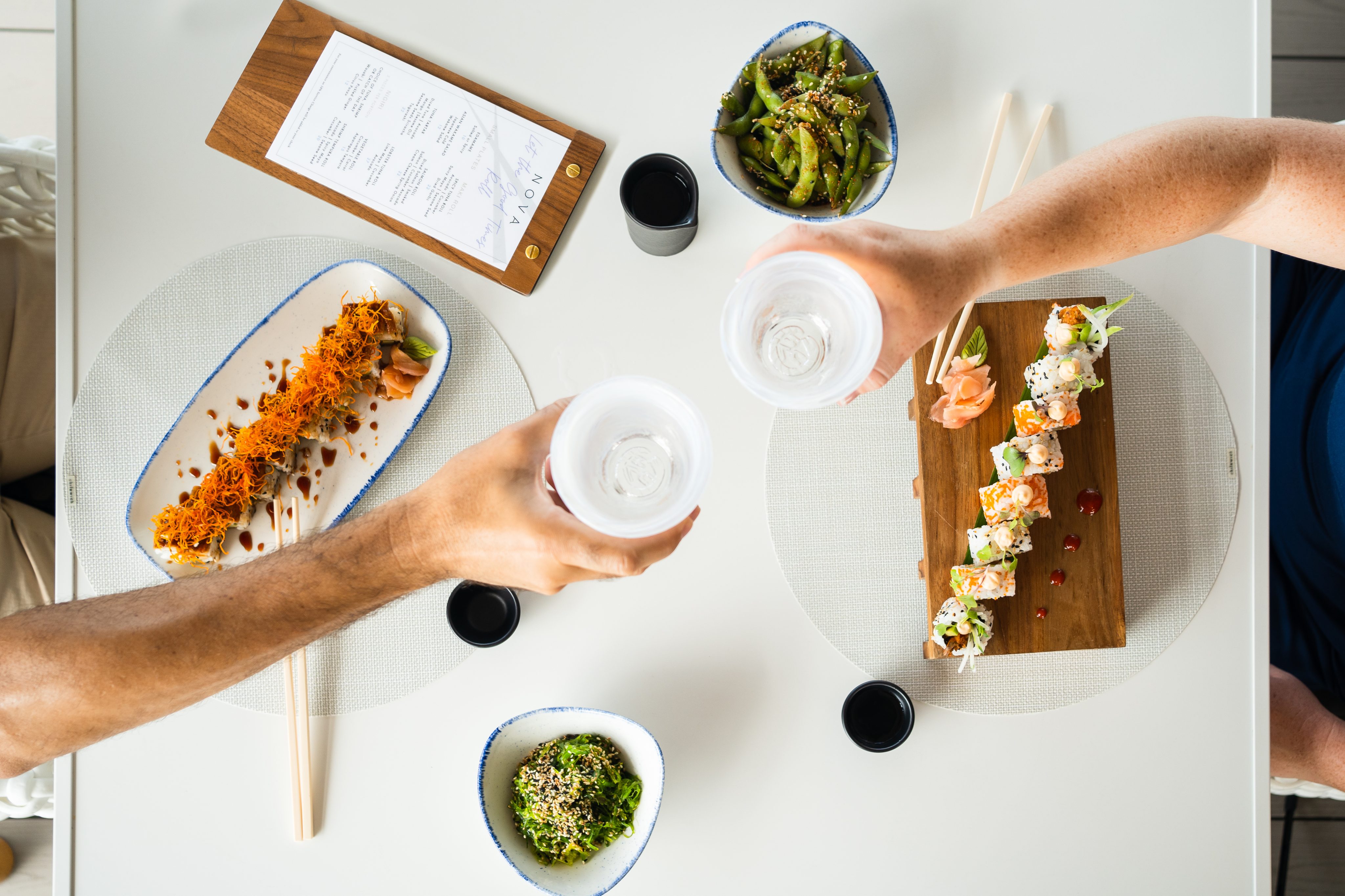 Two people toast with clear glasses at Nova, a sushi restaurant. The table boasts various dishes, including sushi rolls on rectangular plates, a bowl of seaweed salad, and edamame. Chopsticks and a menu grace the white tablecloth.