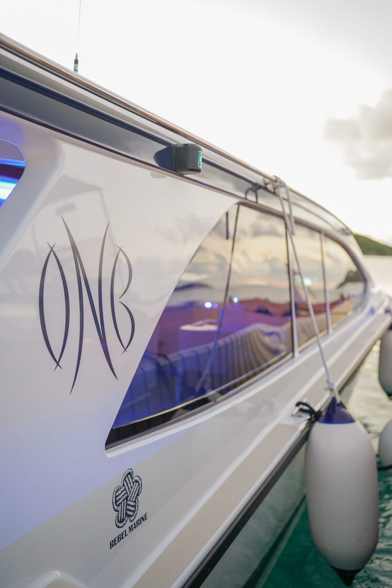 A sleek white boat with shiny windows and the initials "ONB" on its side. The boat, named Odyssey, is adorned with blue ambient lighting and fenders, and is docked in calm water with a blurry shoreline in the background. The logo of "Rebel Marine" is also visible.