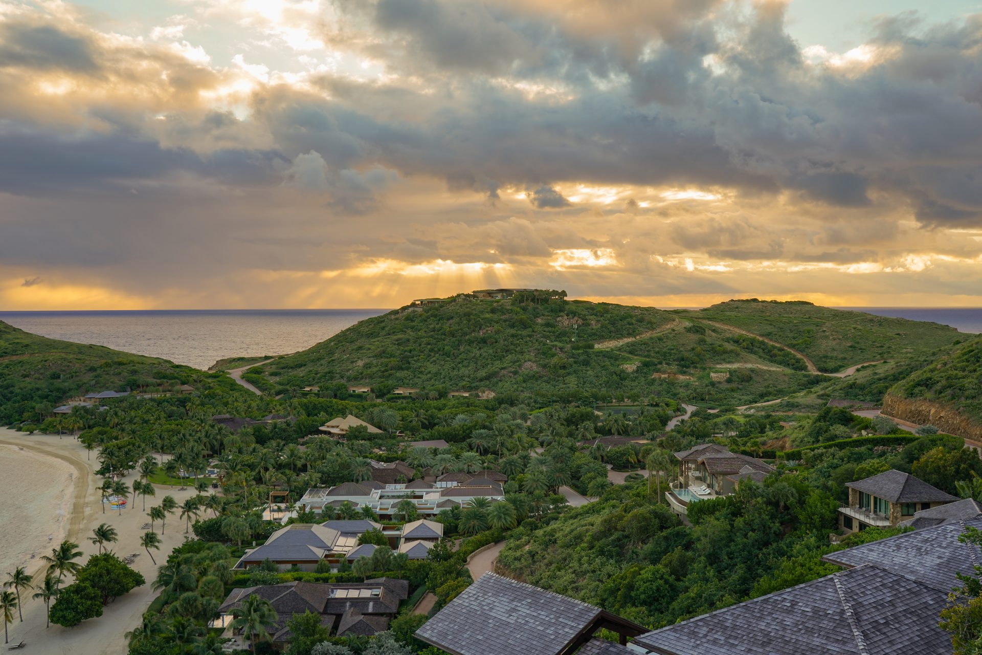 A scenic view of a coastal landscape at sunset. Gentle hills covered with greenery surround a residential area with houses and palm trees. The sky is partly cloudy, sun rays breaking through and casting a golden hue over the ocean and hills, creating a breathtaking spectrum of colors in nature.