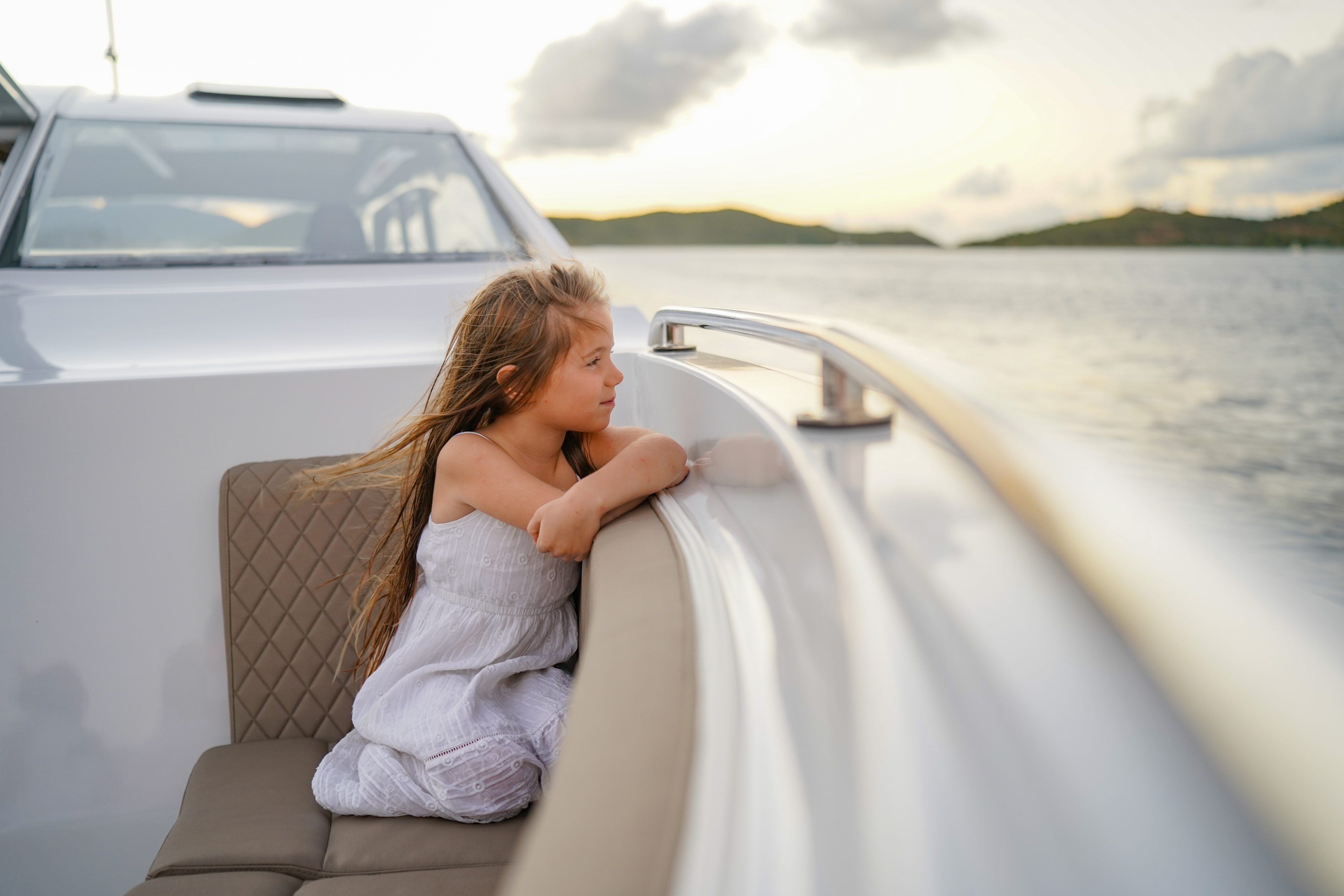 A young girl with long hair gazes thoughtfully over the side of a boat, her arms resting on a railing. She is wearing a white dress and is seated on a cushioned bench. The background shows a calm body of water and distant hills under a cloudy sky—an ideal scene for those places to get married in the BVI.