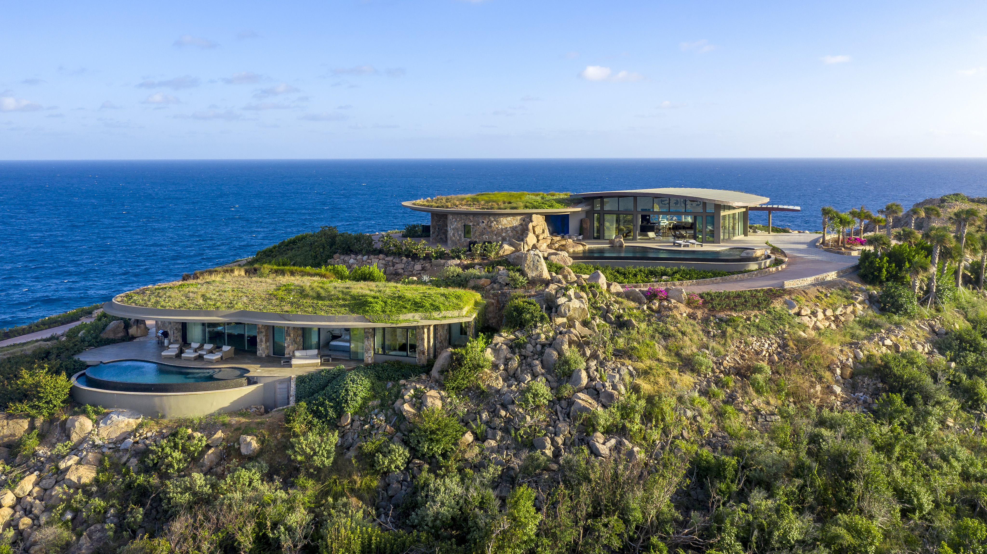 A luxurious, modern clifftop villa, Halo Villa, overlooking the ocean. The villa features multiple levels with large windows, open spaces, and flat roofs adorned with greenery. A pristine infinity pool is visible in the foreground, surrounded by lush vegetation and rocky terrain.