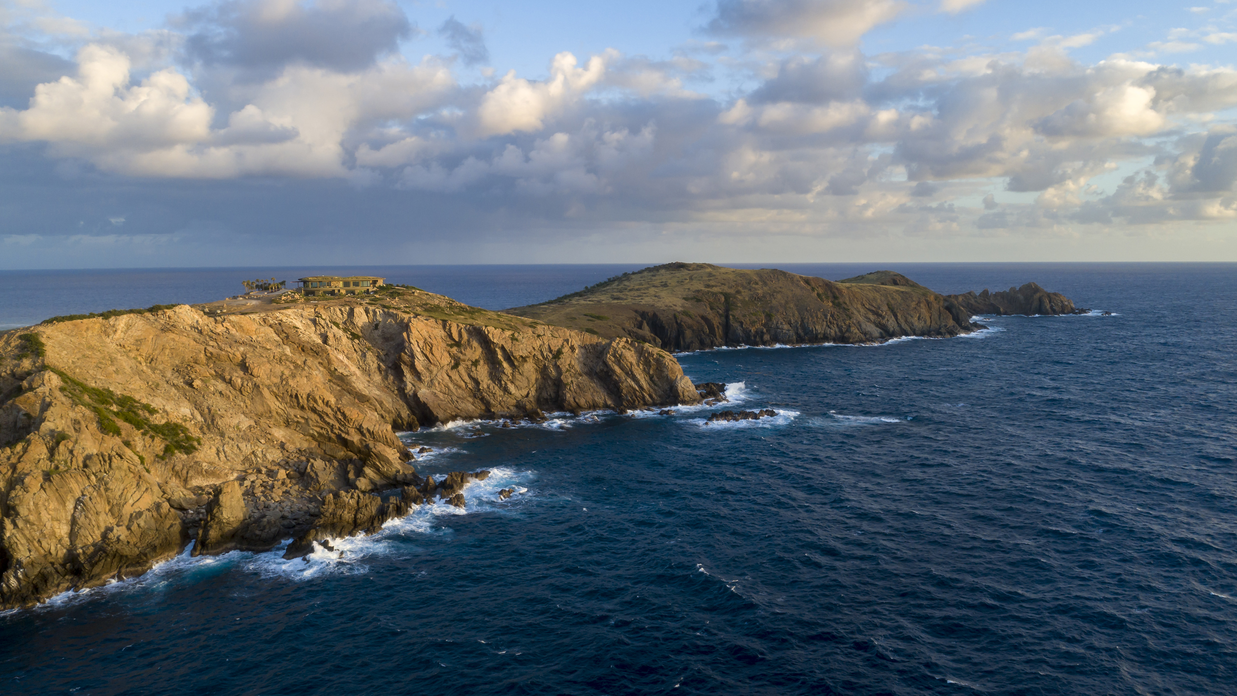 A scenic aerial view of a rugged coastline with cliffs meeting the ocean reveals Halo Villa perched near the edge of the cliffs on the left. The sky is partly cloudy, and the sea below is a deep blue with gentle waves crashing against the rocks.