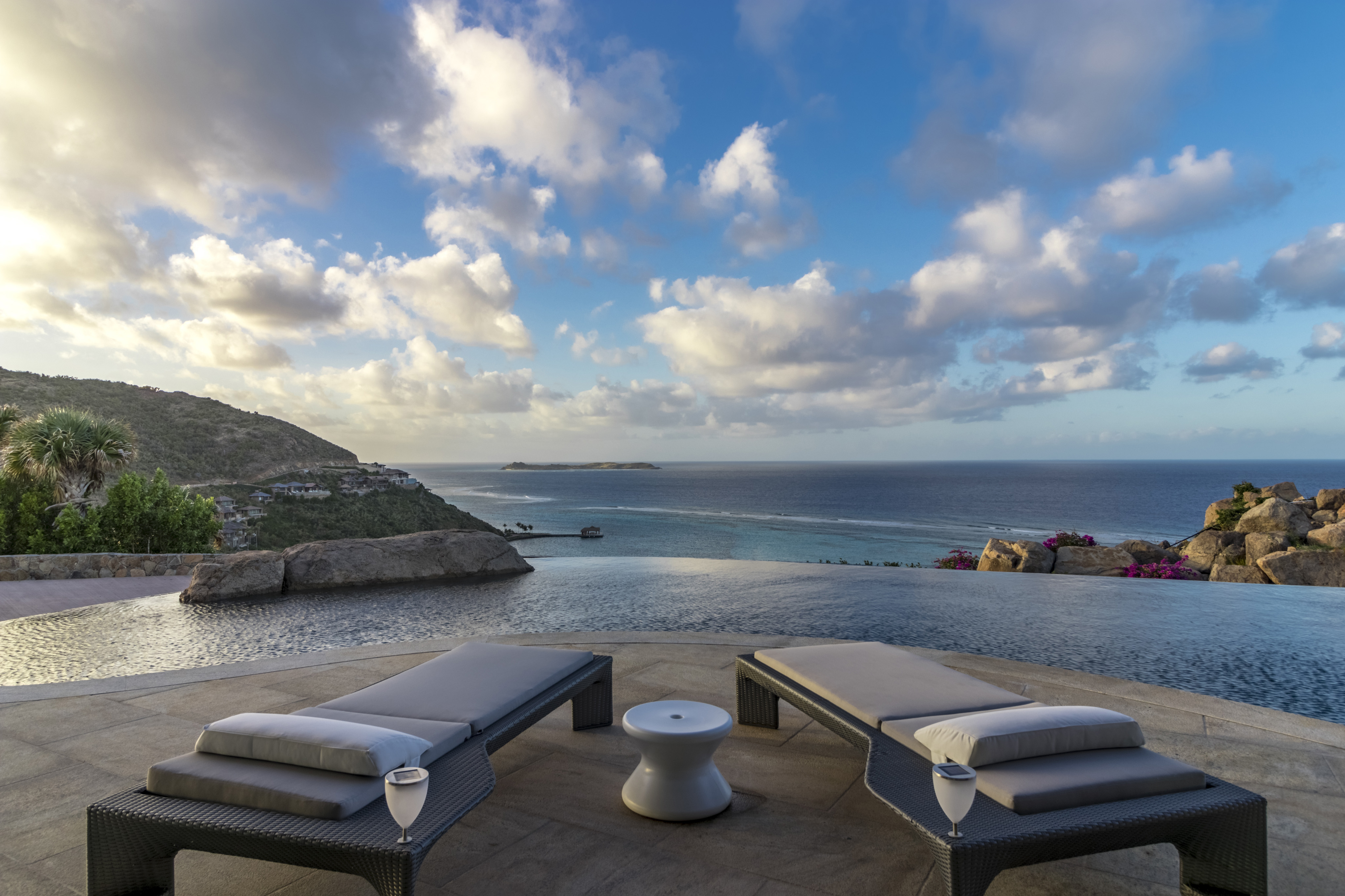 An infinity pool at the villa overlooks a scenic view of the ocean and distant islands under a partly cloudy sky. Two lounge chairs and a small table rest on the pool deck, with glasses placed on the table. Rocky hillsides and foliage frame the tranquil scene, creating an idyllic halo of serenity.