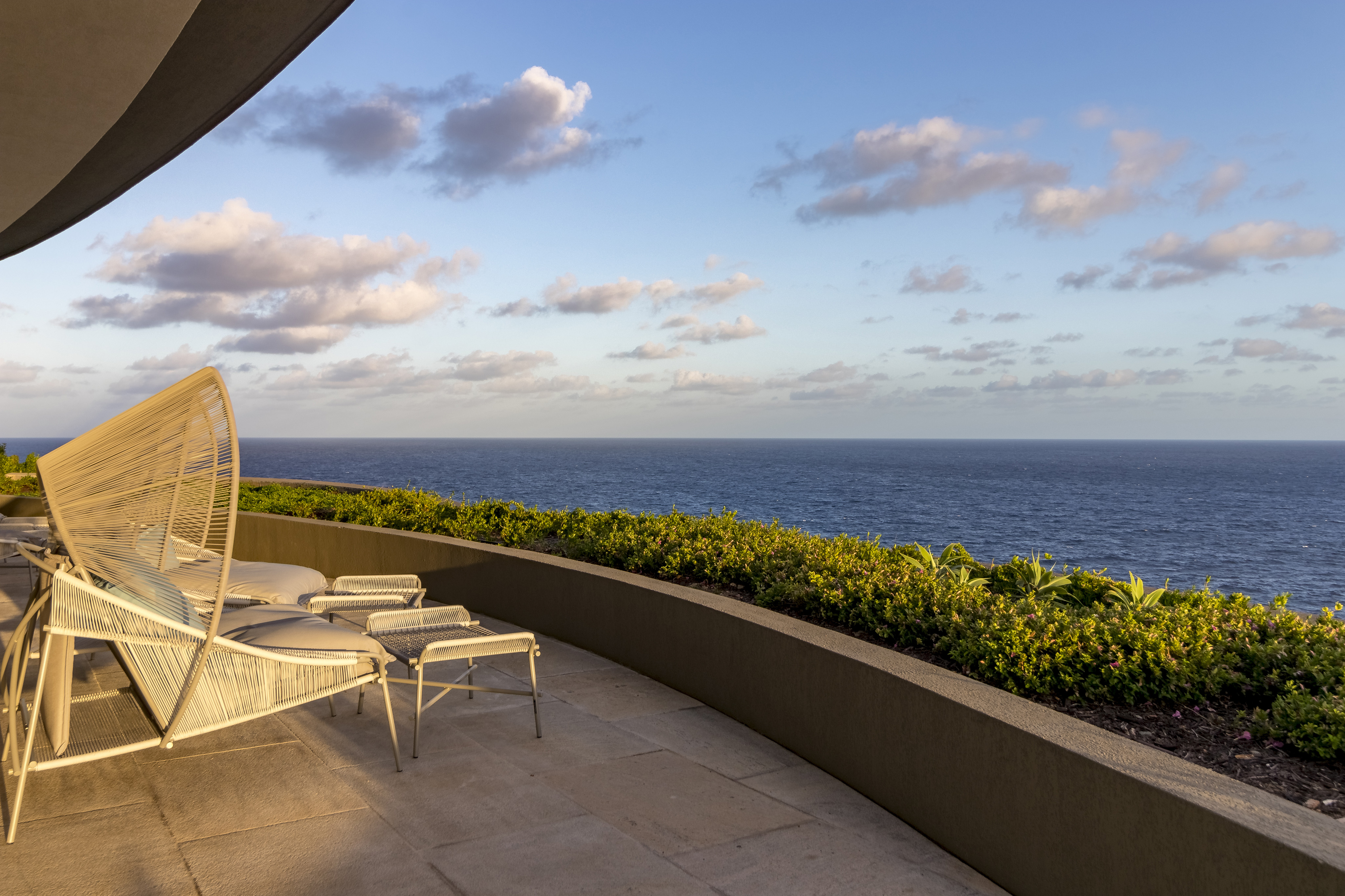 Outdoor patio at Halo Villa with modern white loungers and a canopy, overlooking a vast ocean view under a partly cloudy sky. The patio features a low, curved retaining wall with lush greenery along the edge, framing the scenic seascape.