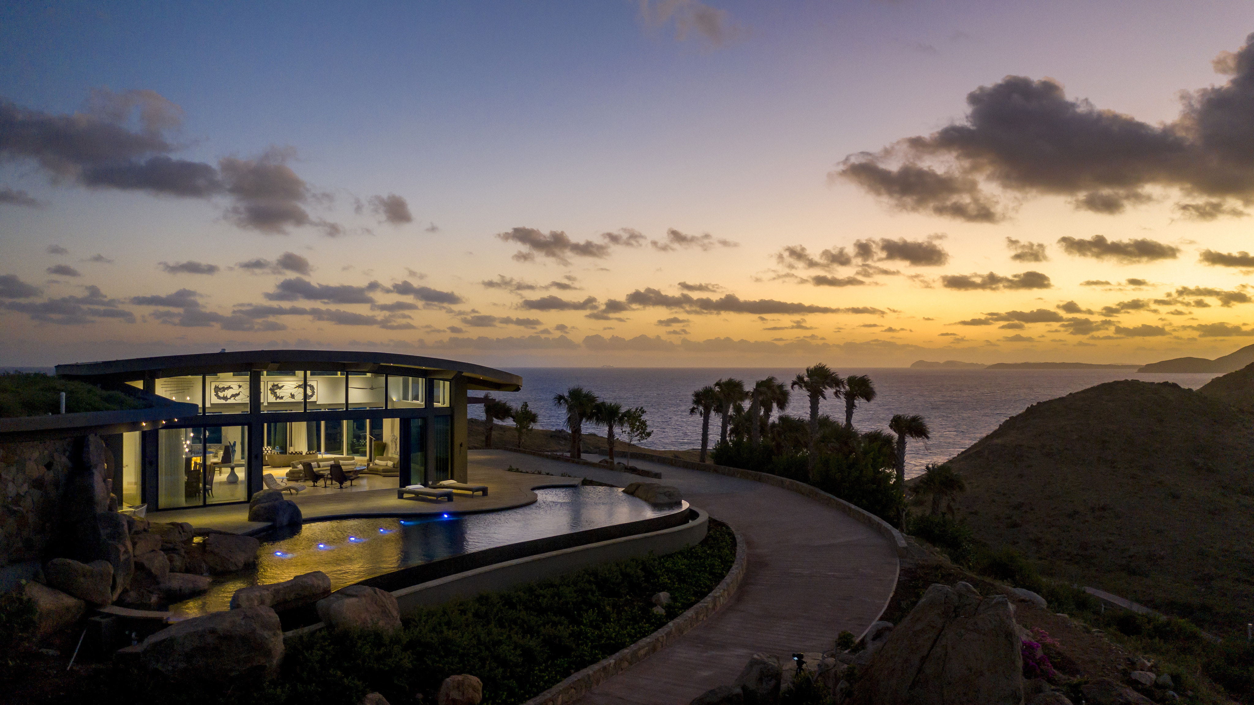 Halo Villa, a modern glass-walled house, is situated on a coastal hill overlooking the ocean at sunset. The villa features an illuminated swimming pool, palm trees, and a winding path leading up to it. The sky is adorned with scattered clouds, creating a serene and picturesque scene.