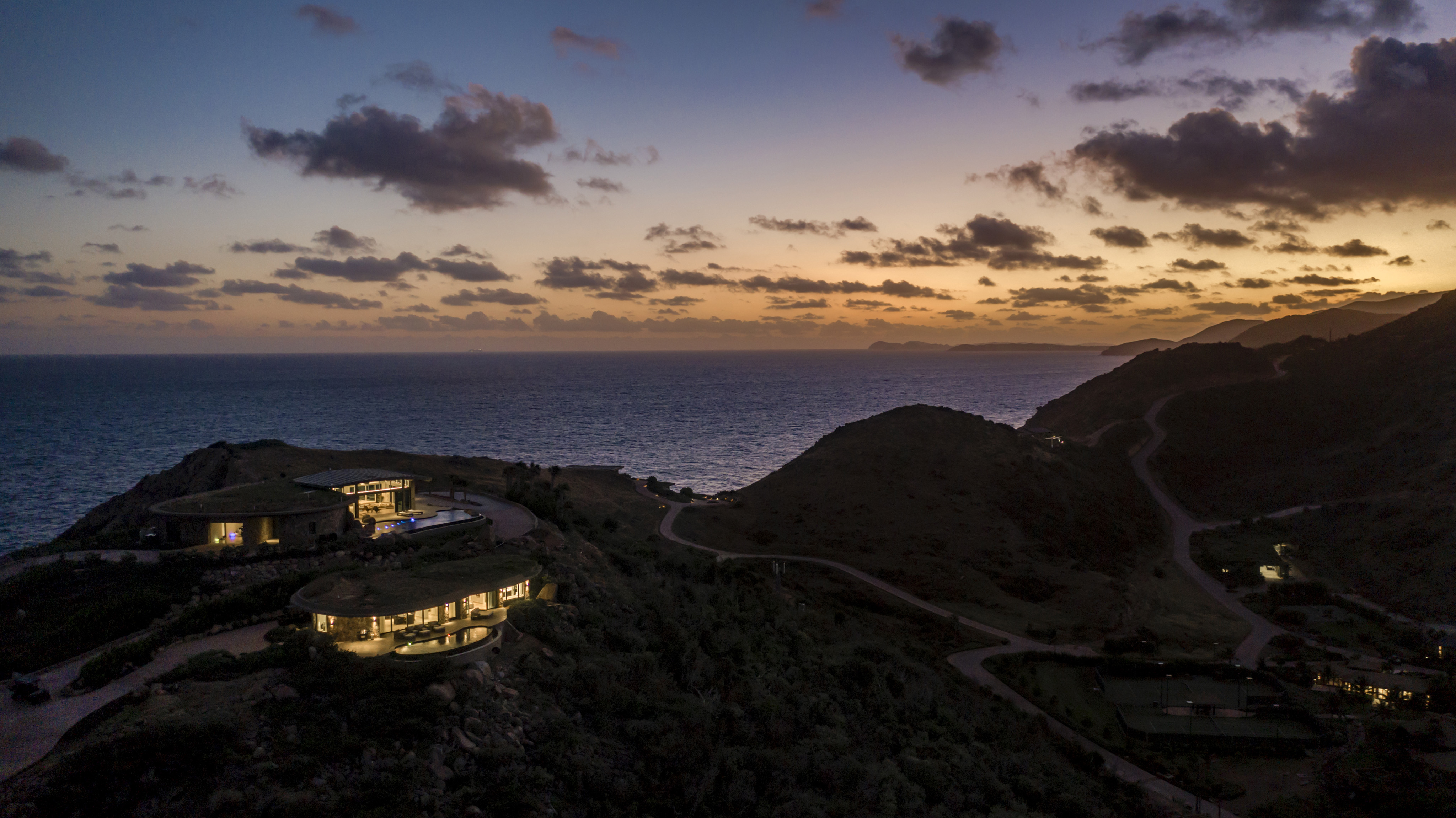 A scenic view of a coastal landscape at dusk, featuring modern villas on hills overlooking the ocean. The sky is painted with hues of orange and purple as the sun sets, and the lights from the houses cast a warm halo. Winding roads can be seen among the hills.