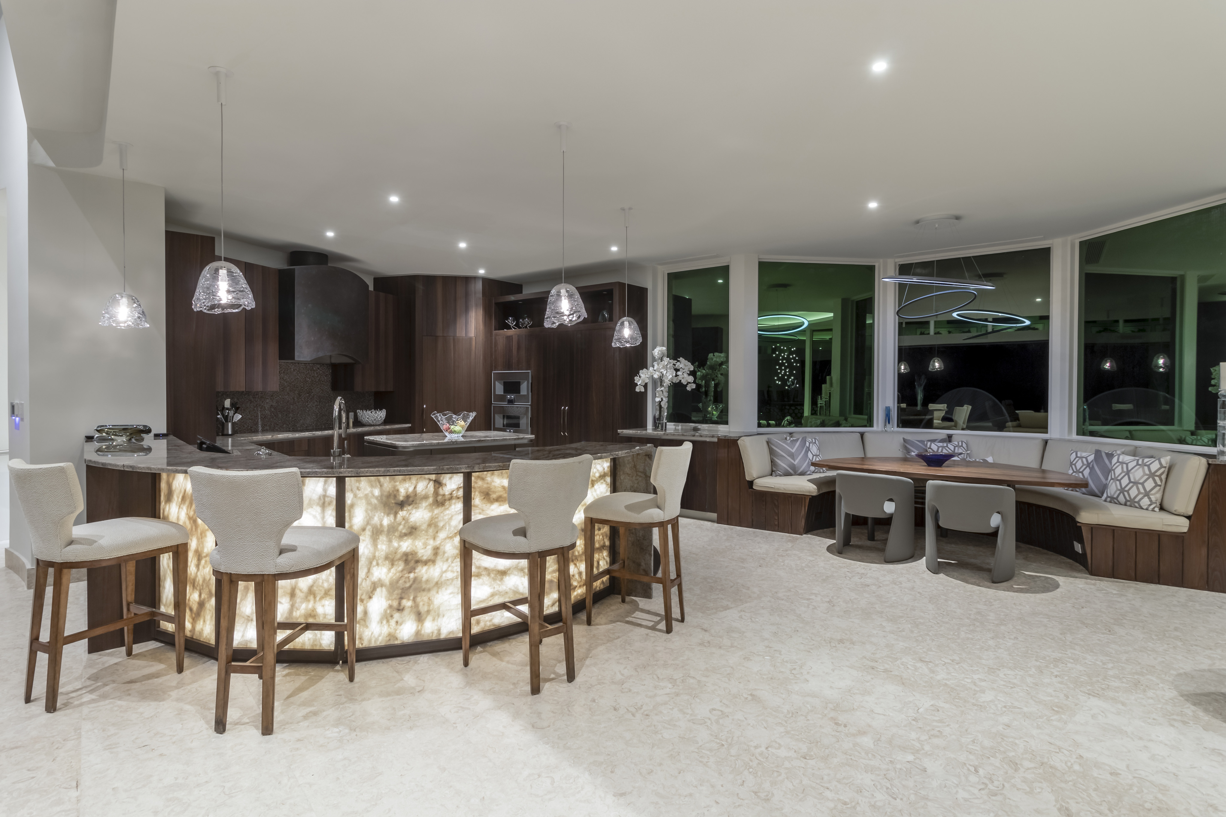 Modern kitchen at Halo Villa with a central island featuring illuminated panels and bar stools. Pendant lights hang above the island. The kitchen boasts dark cabinetry, built-in appliances, and a cozy dining nook with cushioned seating in front of large windows.