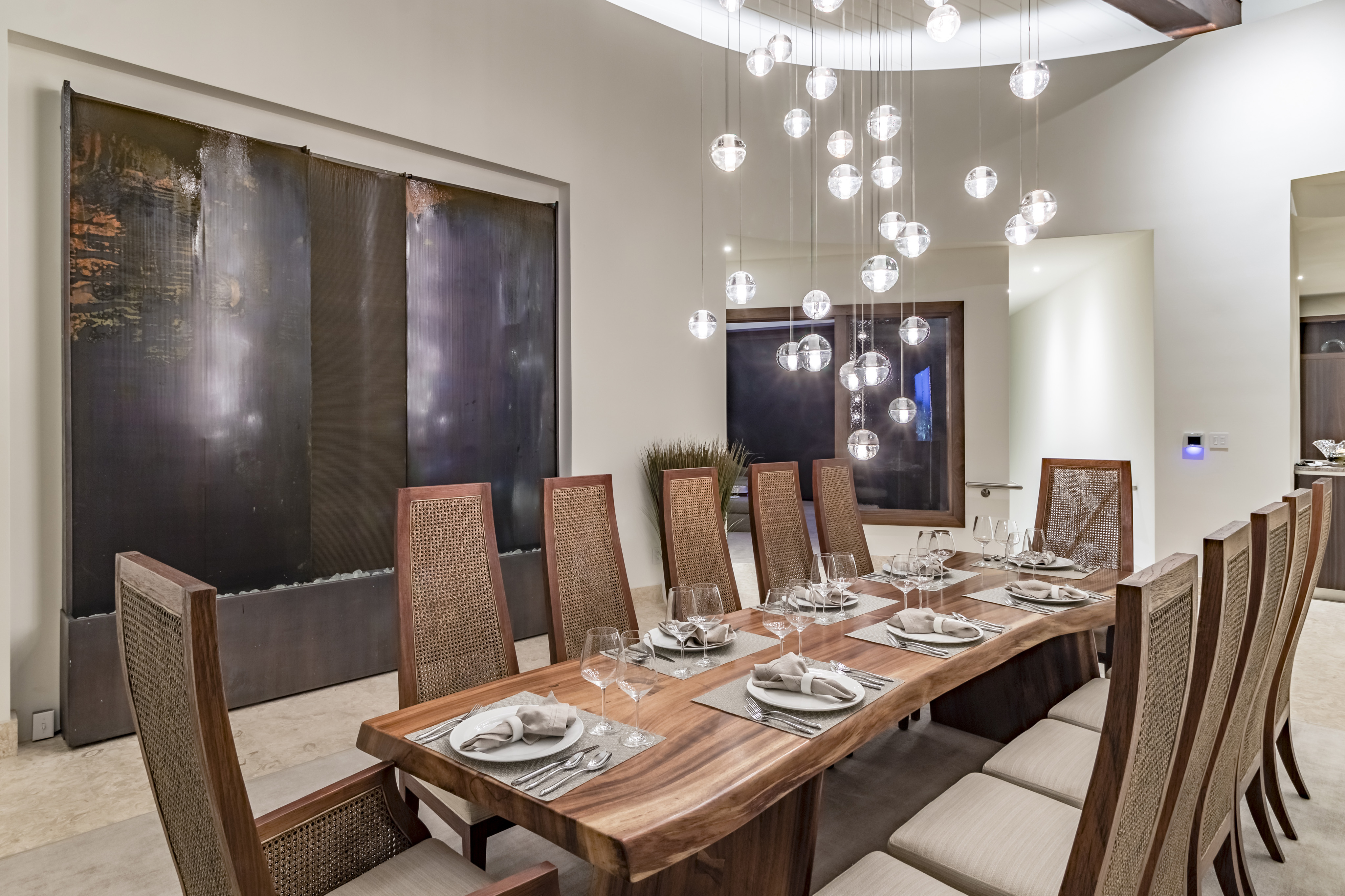 A modern villa dining room features a long wooden table with ten cushioned chairs placed evenly around it. The table is set with plates, napkins, and glasses. Above, multiple halo pendant lights hang from the ceiling. In the background, textured wall panels add decorative flair.