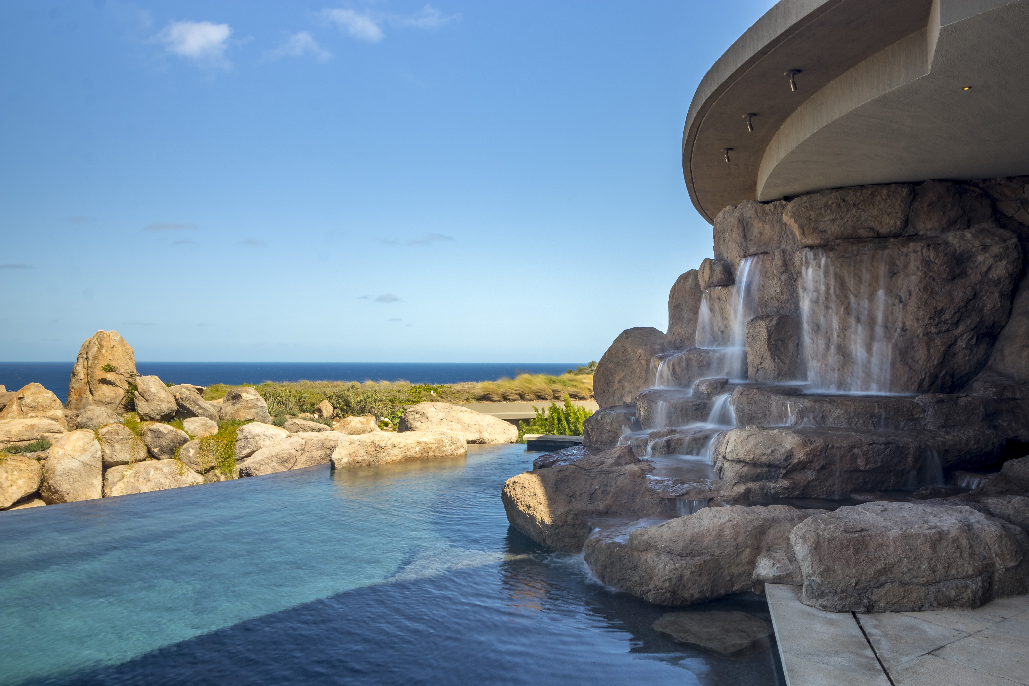 Halo Villa's luxurious infinity pool overlooks a serene ocean view. The pool's edge blends seamlessly with the horizon, and a cascading waterfall flows from a rocky structure on the right, creating a tranquil atmosphere. Large rocks and foliage enhance the natural aesthetic of this exquisite retreat.