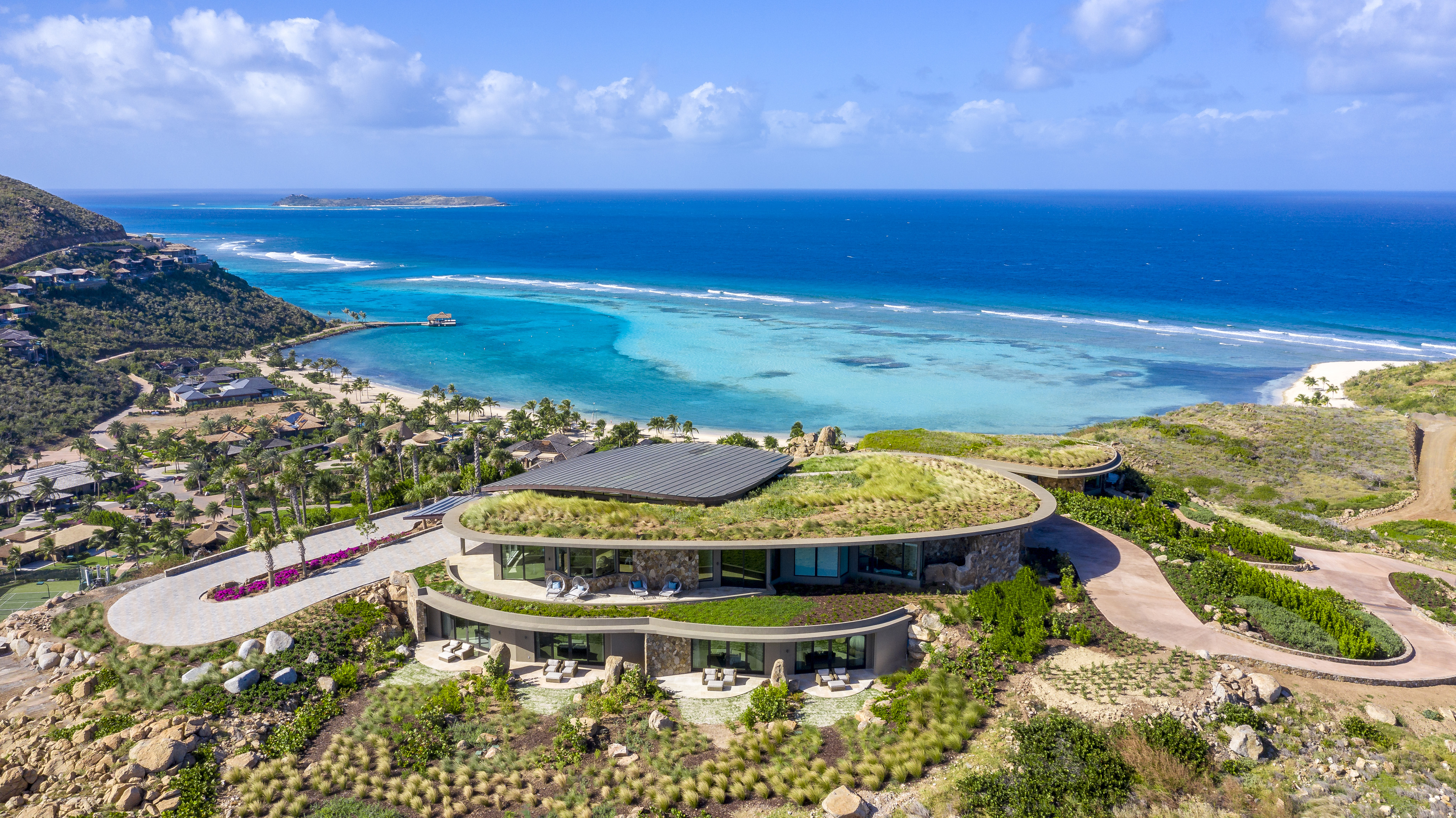 Halo Villa is a modern, multi-level house with a green rooftop that overlooks a beautiful, expansive turquoise ocean. The surrounding landscape includes rocky hills, lush vegetation, and nearby houses along the coastline, all under a partly cloudy sky.