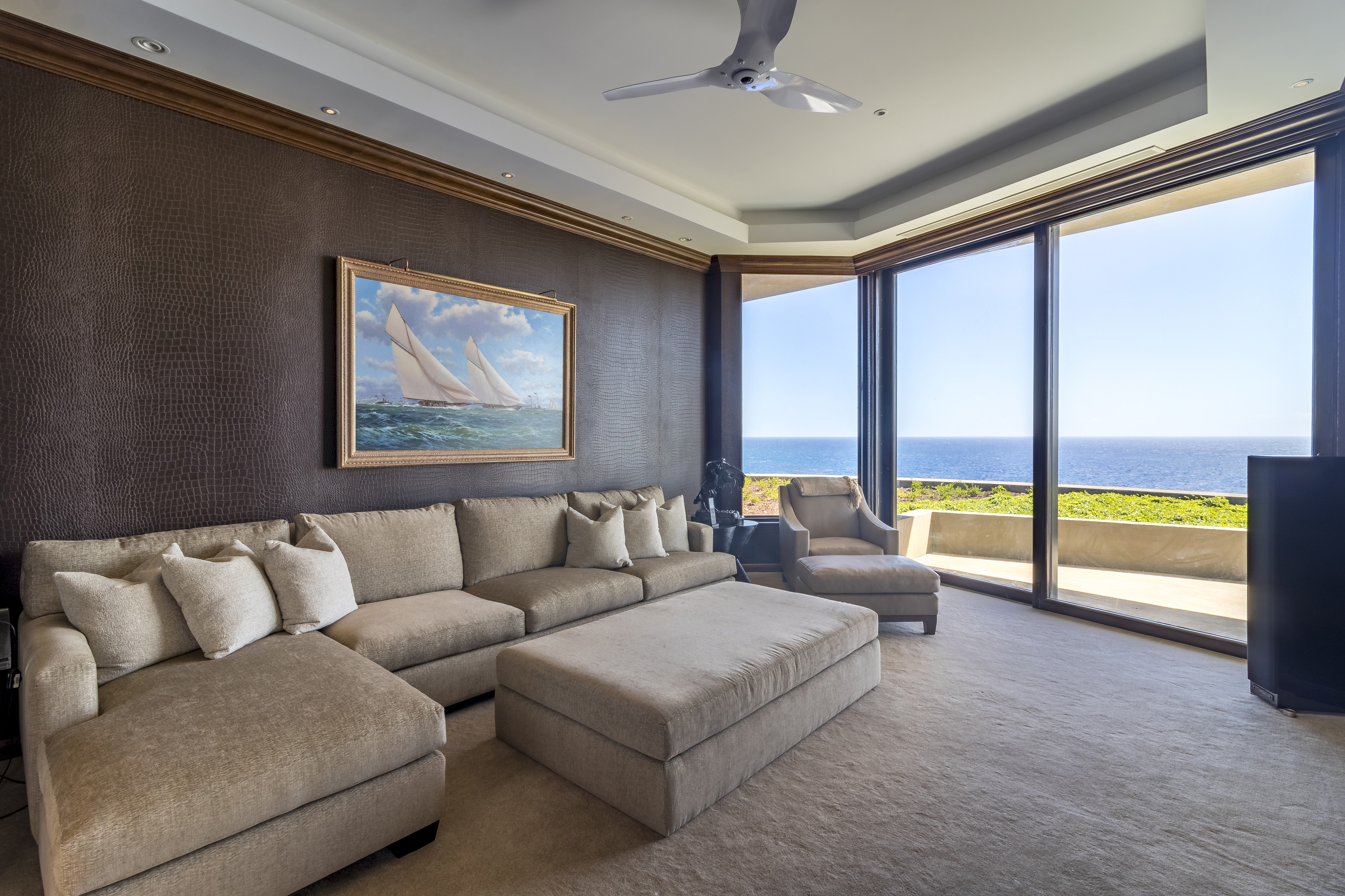 A modern living room in Halo Villa features a large beige sectional sofa, matching ottoman, and pillows against a dark textured wall. A framed painting of sailboats hangs above the sofa. Floor-to-ceiling windows reveal an expansive ocean view and a balcony with a glass railing.