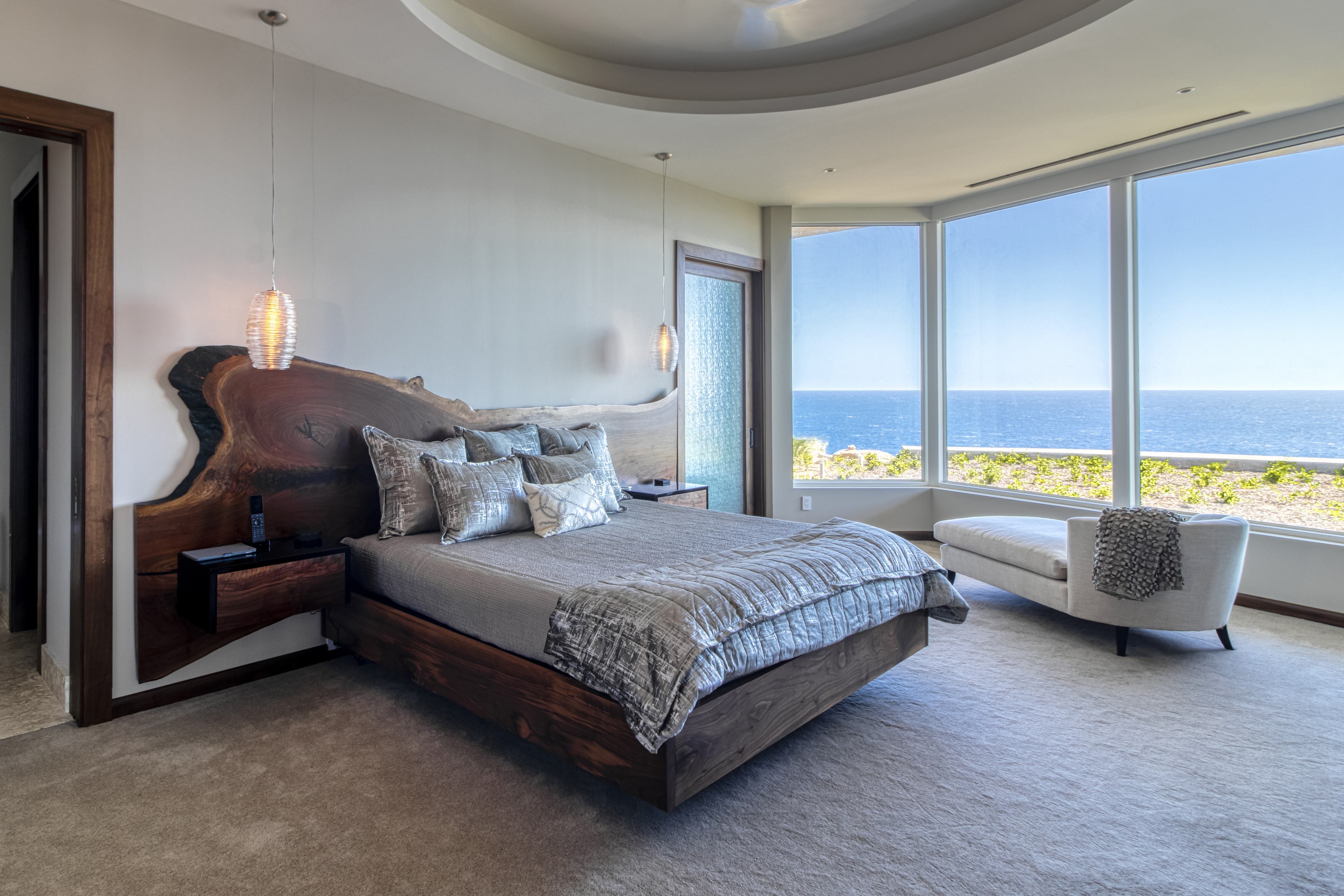 A modern bedroom at Halo Villa, featuring a large wooden bed upholstered in grey bedding and pillows with a natural wood headboard. The room boasts grey carpet, pendant lights, a chaise lounge by floor-to-ceiling windows revealing an ocean view, and a frosted glass door on the left wall.