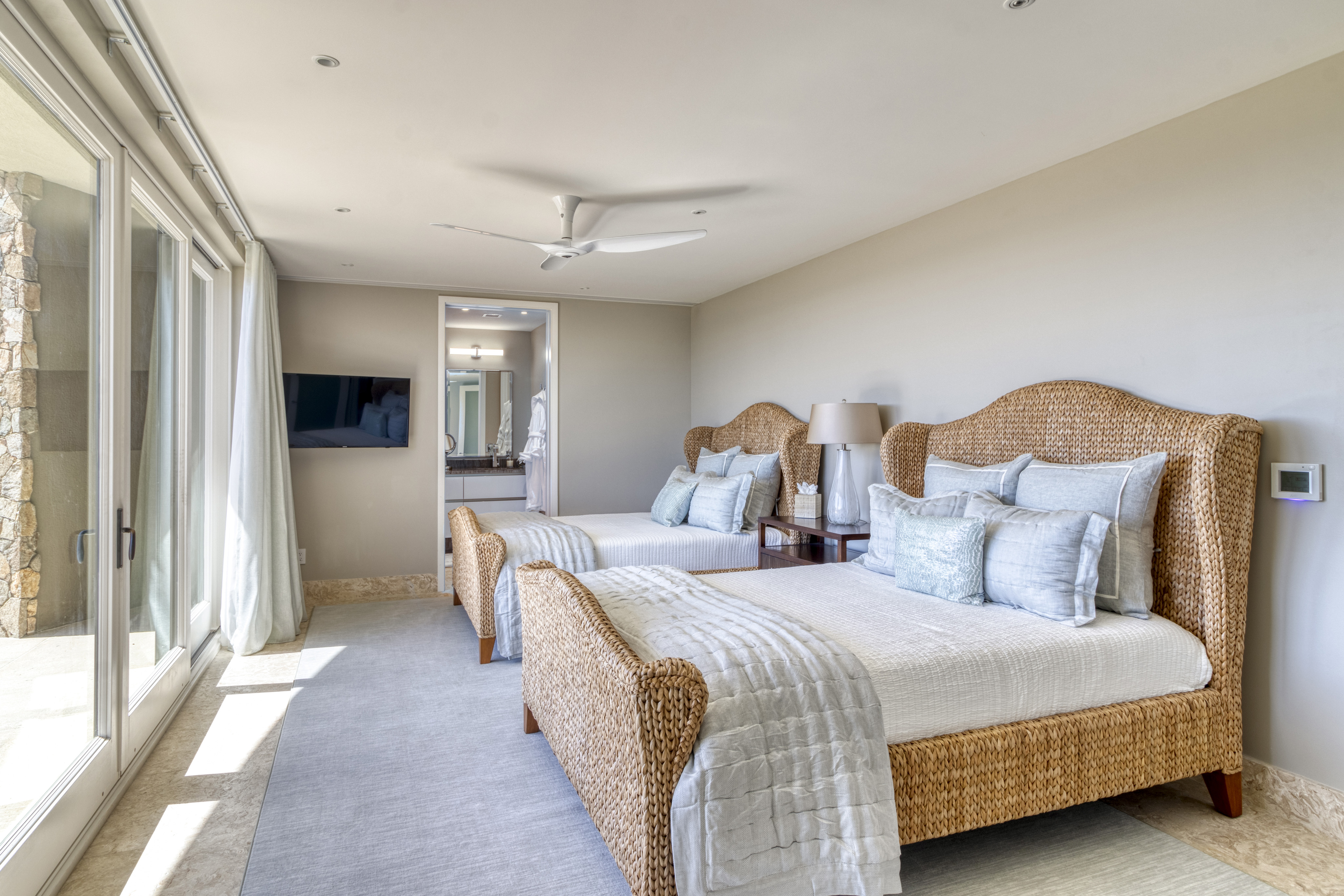 A serene bedroom in the villa features two wicker beds with white linens and pastel blue cushions. A nightstand with a halo lamp is placed between the beds. The room has floor-to-ceiling windows, a flat-screen TV on the wall, and a view into the adjoining bathroom.