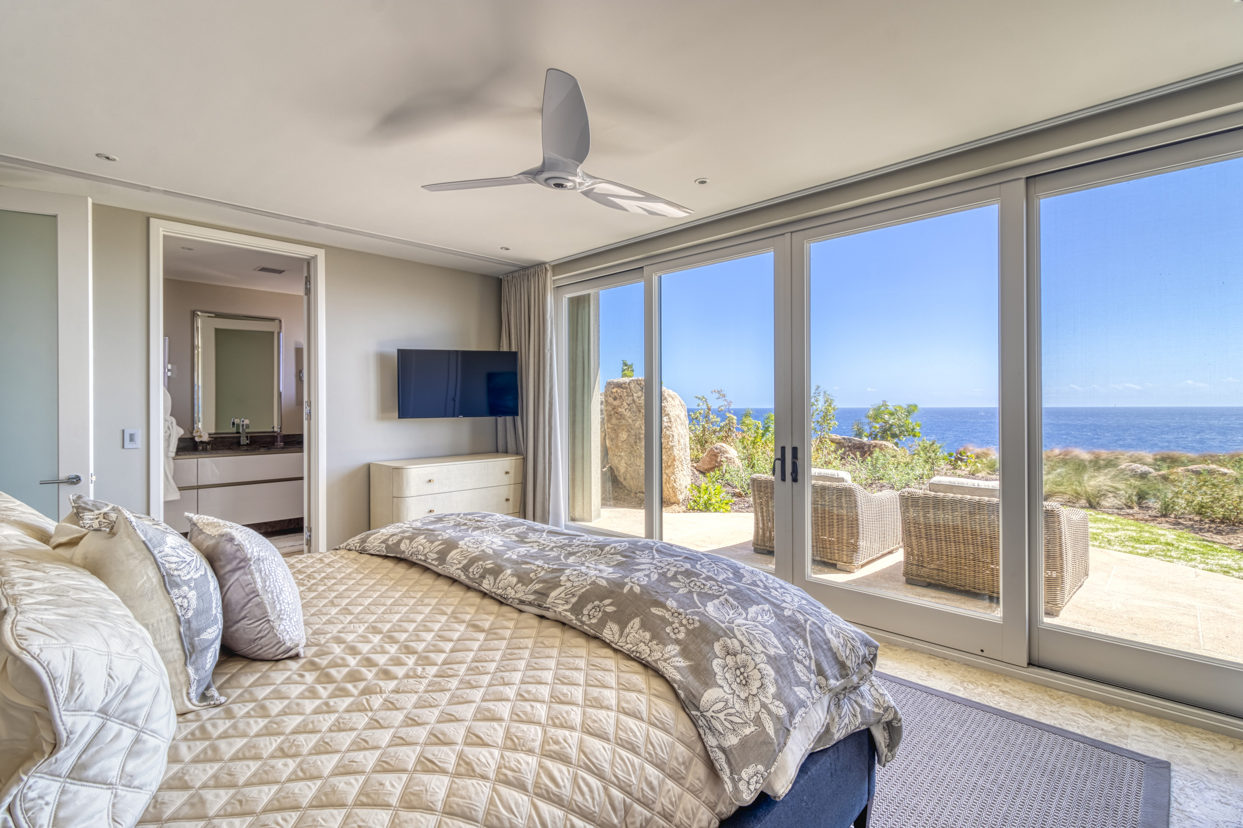 A modern bedroom in Halo Villa features a king-sized bed adorned with a patterned beige comforter and pillows. Floor-to-ceiling windows offer a panoramic ocean view, with a patio featuring outdoor furniture. A ceiling fan, wall-mounted TV, and dresser complete the room's decor.