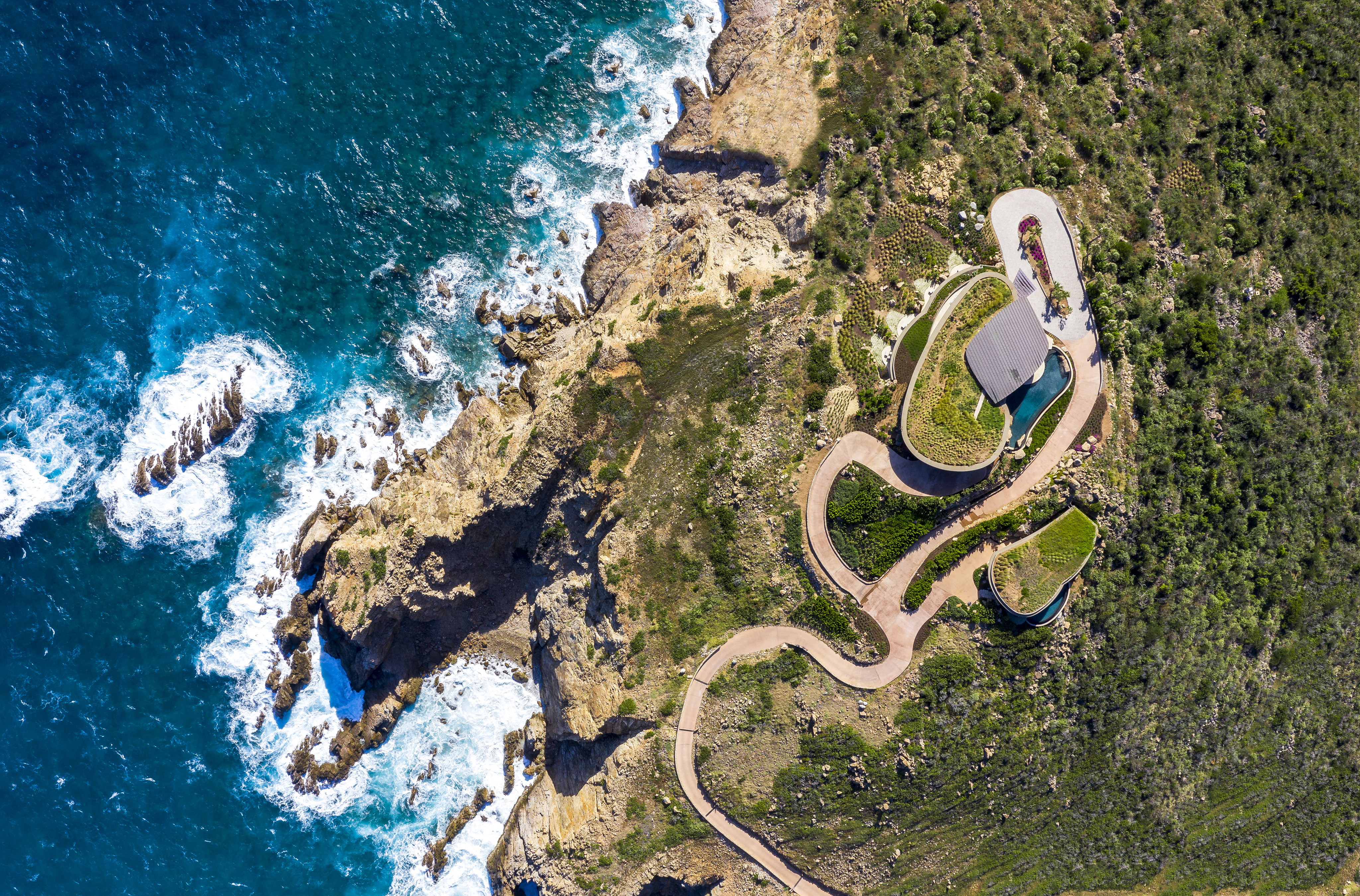 Aerial view of a coastline with rugged cliffs and crashing waves. A winding pathway leads to a modern villa with a green roof, resembling a halo in the lush, green landscape. The surrounding scenery contrasts beautifully with the deep blue ocean.