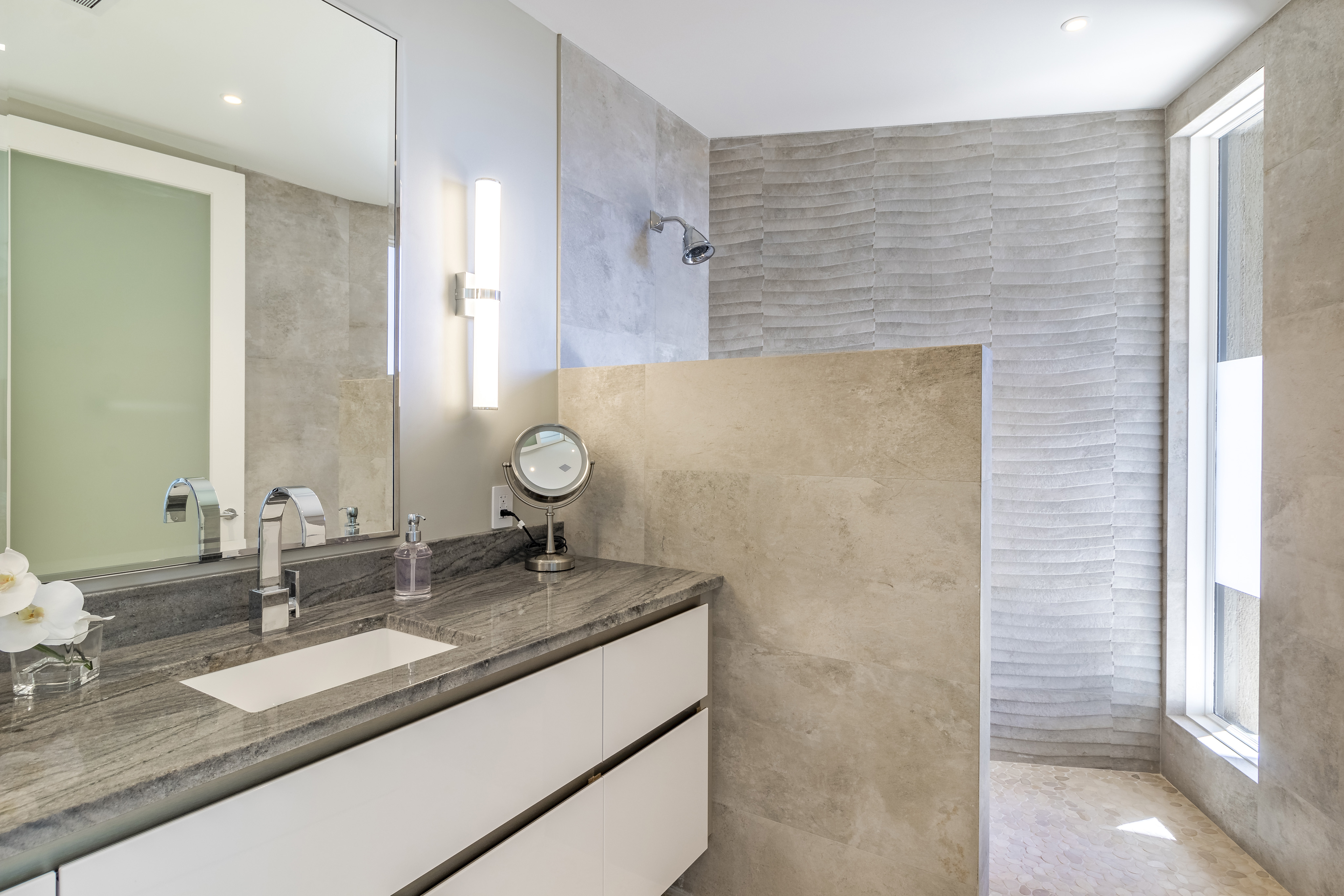 The Halo Villa's modern bathroom boasts a large stone-topped vanity with dual sinks and sleek faucets. A lighted mirror hangs on the wall above, complemented by an additional circular mirror on the counter. The walk-in shower features a textured stone wall and glass partition, adding to the luxurious feel.