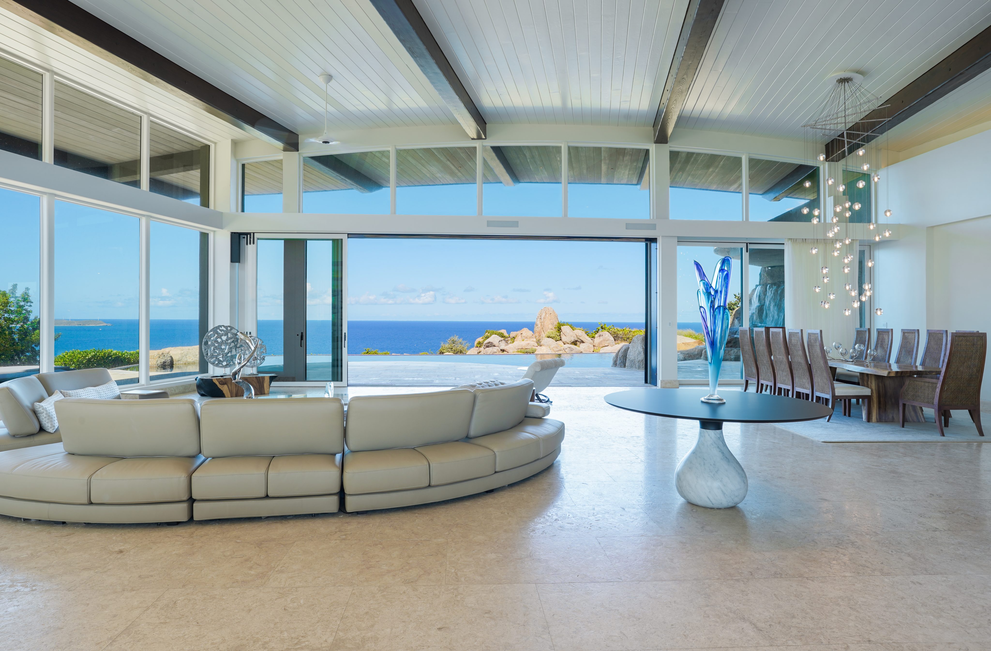 Welcome to Halo Villa's spacious modern living area with large windows offering an ocean view. It features a curved beige sofa, round coffee table, and a long dining table with multiple chairs. High ceilings with exposed beams and a contemporary chandelier accentuate the room's elegance.