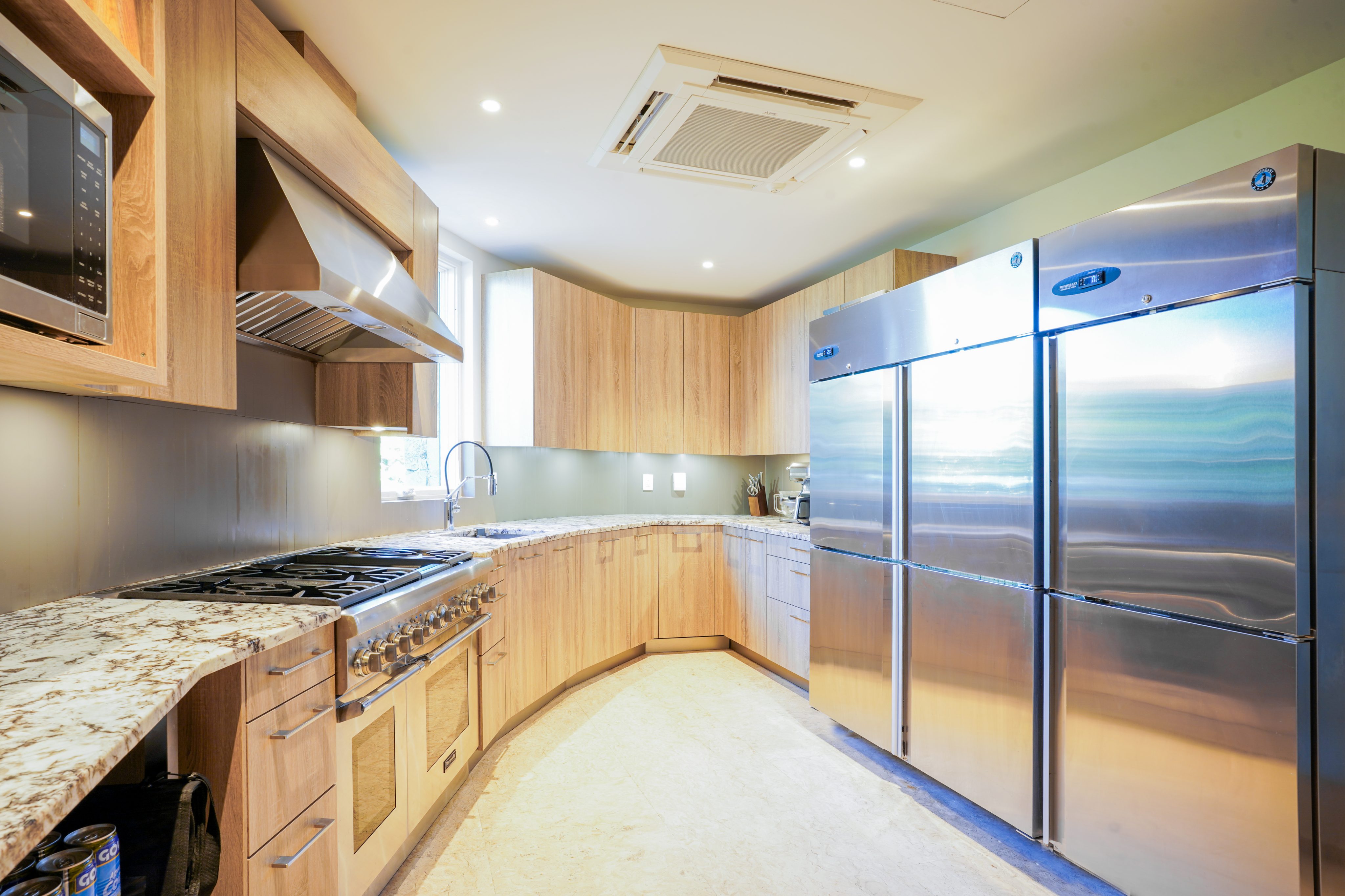 A modern kitchen at Halo Villa featuring wooden cabinets, a stainless steel range with a ventilation hood, a built-in microwave, and a large double-door stainless steel refrigerator. The marble countertops glisten under the recessed lighting on the ceiling.