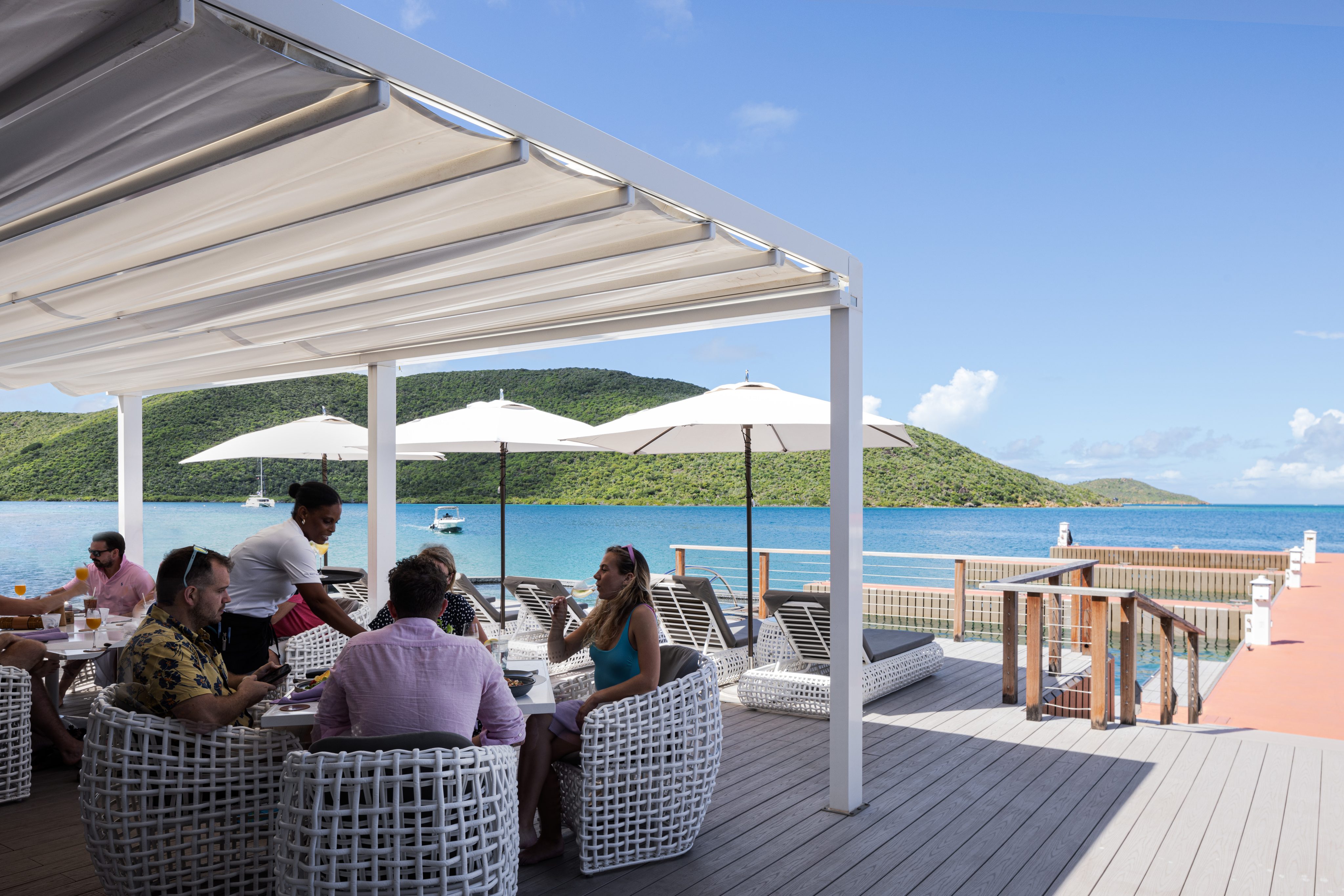 People are seated at a shaded outdoor patio with a white awning, enjoying a meal beside a clear blue body of water. Sun umbrellas stand near the patio, and green hills are visible in the background. A server is attending to the people at the table, ensuring they have everything for a pleasant stay.