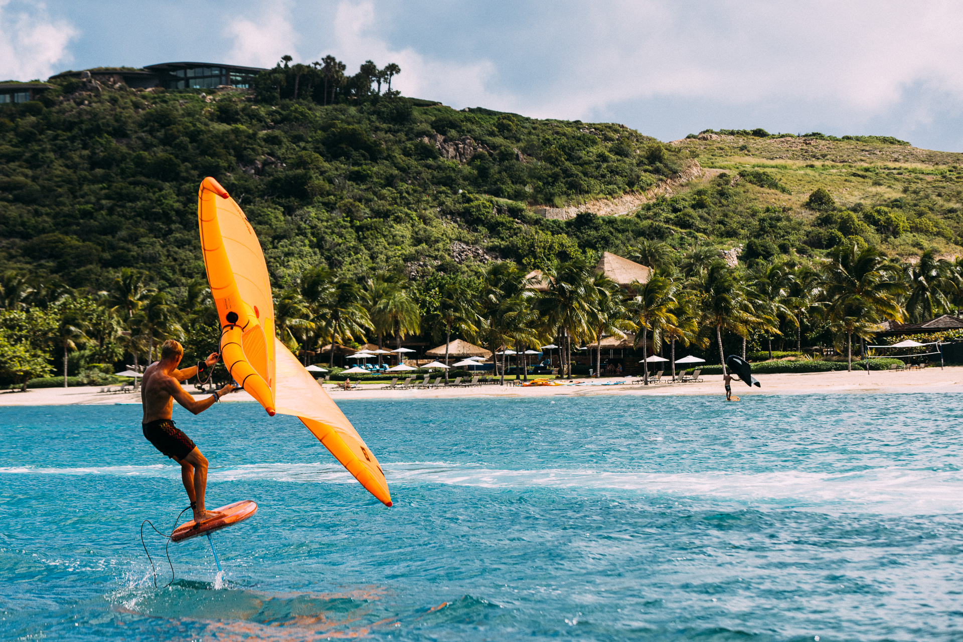 A person is seen surfing on a hydrofoil board with an orange wing sail on a turquoise body of water near a tropical beach. The shoreline, where visitors love to stay, is lined with palm trees. In the background, a green hill with buildings adds charm as another surfer rides in the distance.