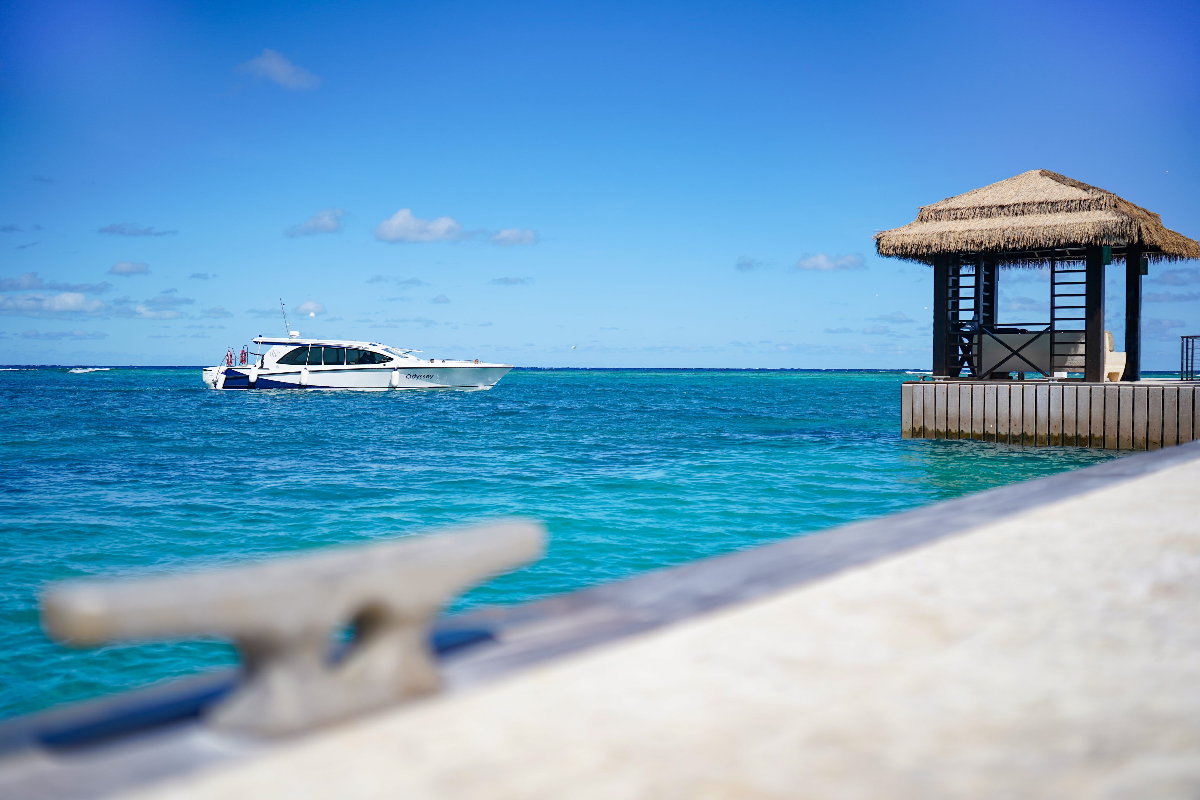 A boat floats on clear blue water under a sunny sky, with a small hut with a thatched roof on the right side of the image. A blurred foreground object, likely part of a dock or waterfront structure, adds to the tranquil scene, inviting you to stay and enjoy this idyllic paradise.