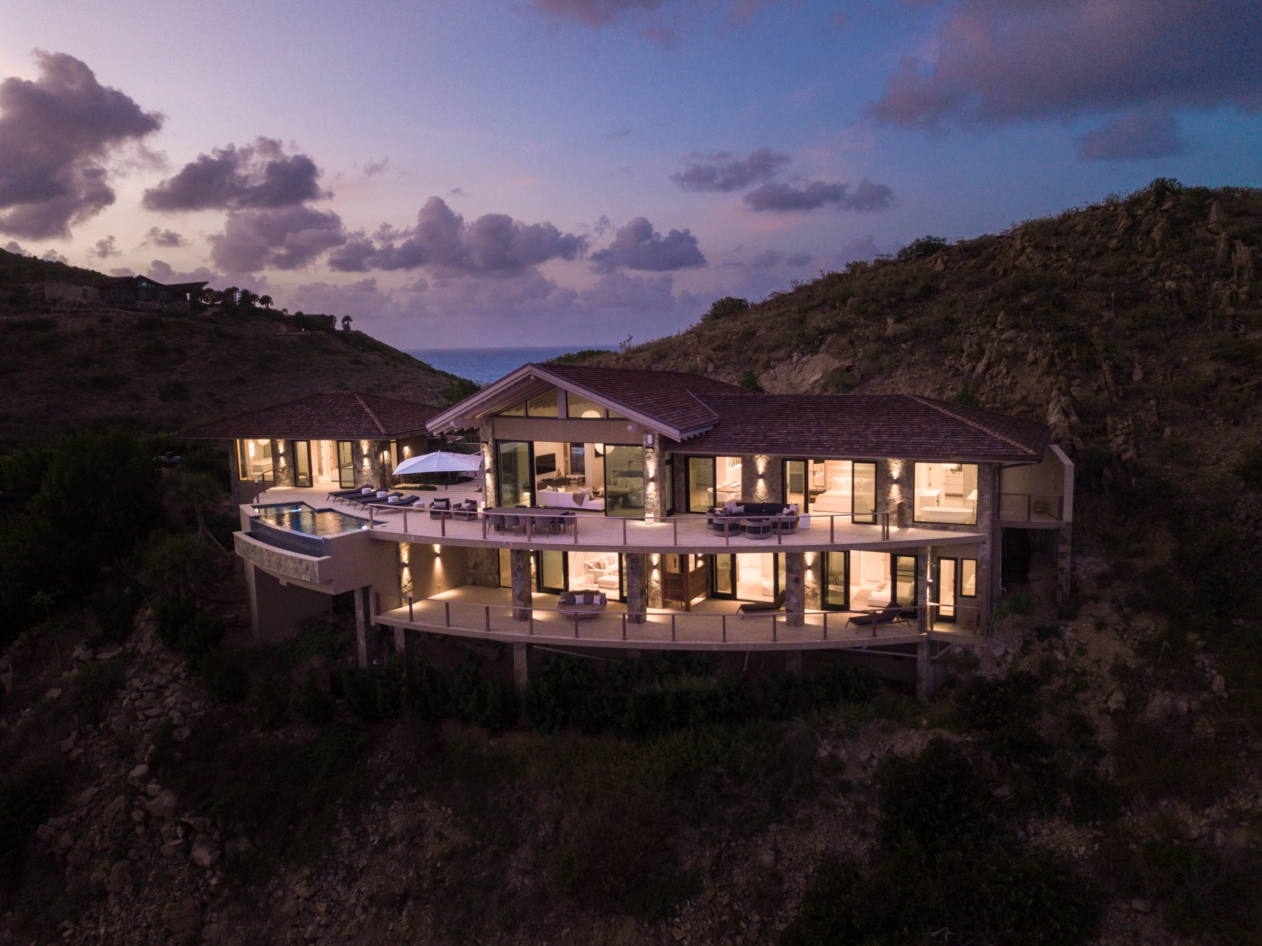 Hummingbird Villa, a luxurious hillside mansion with large windows and multiple balconies, is illuminated at dusk. The house features modern architecture and overlooks a scenic view with distant ocean and cloud-filled sky. This exquisite villa rental blends seamlessly with the surrounding natural landscape.