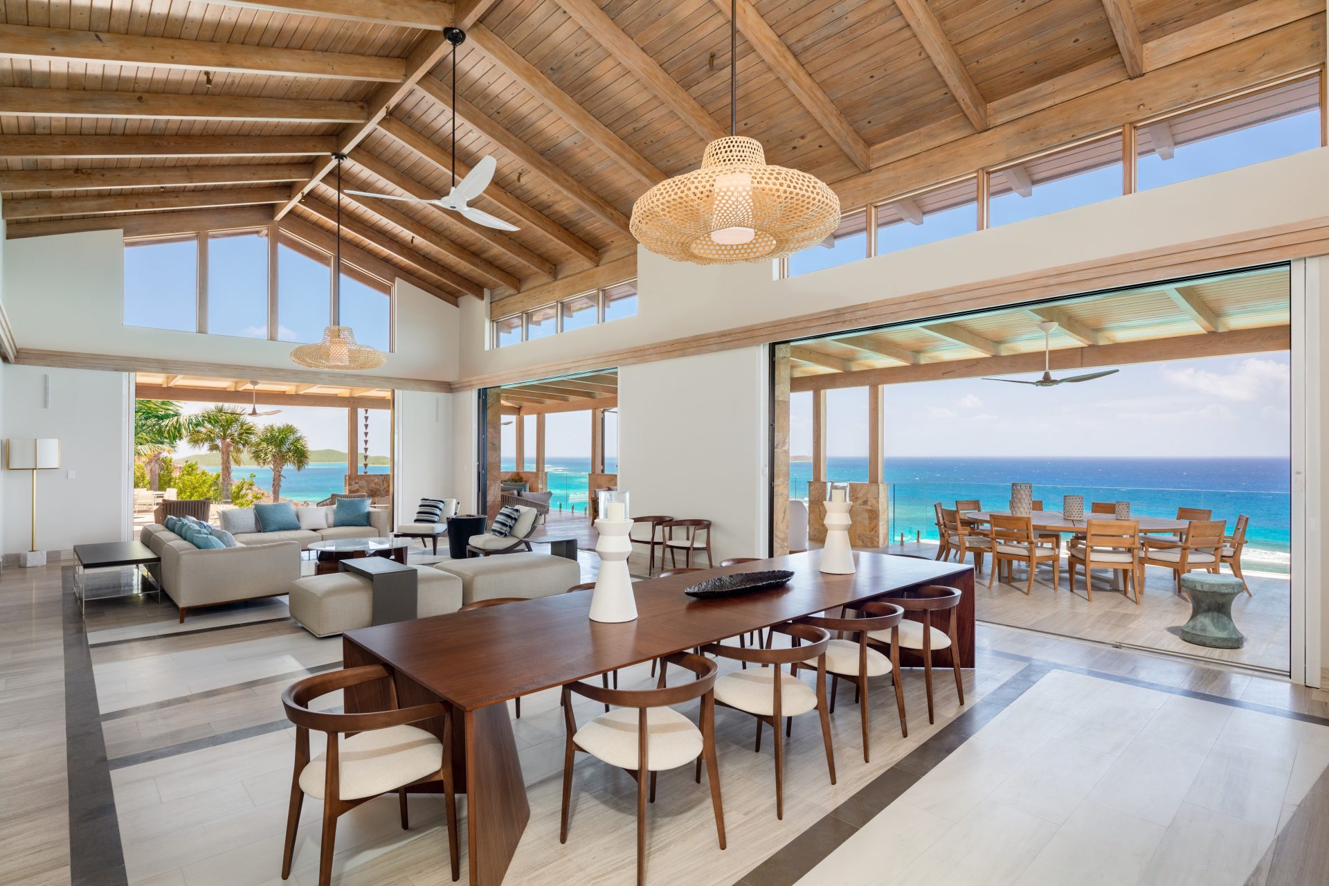 Open-plan luxury Maronti beach villa with high wooden ceilings, modern furniture, and large windows. The dining area features a wooden table with white chairs, while the adjoining living space has plush sofas. A sliding glass door opens to an oceanfront deck with outdoor dining.