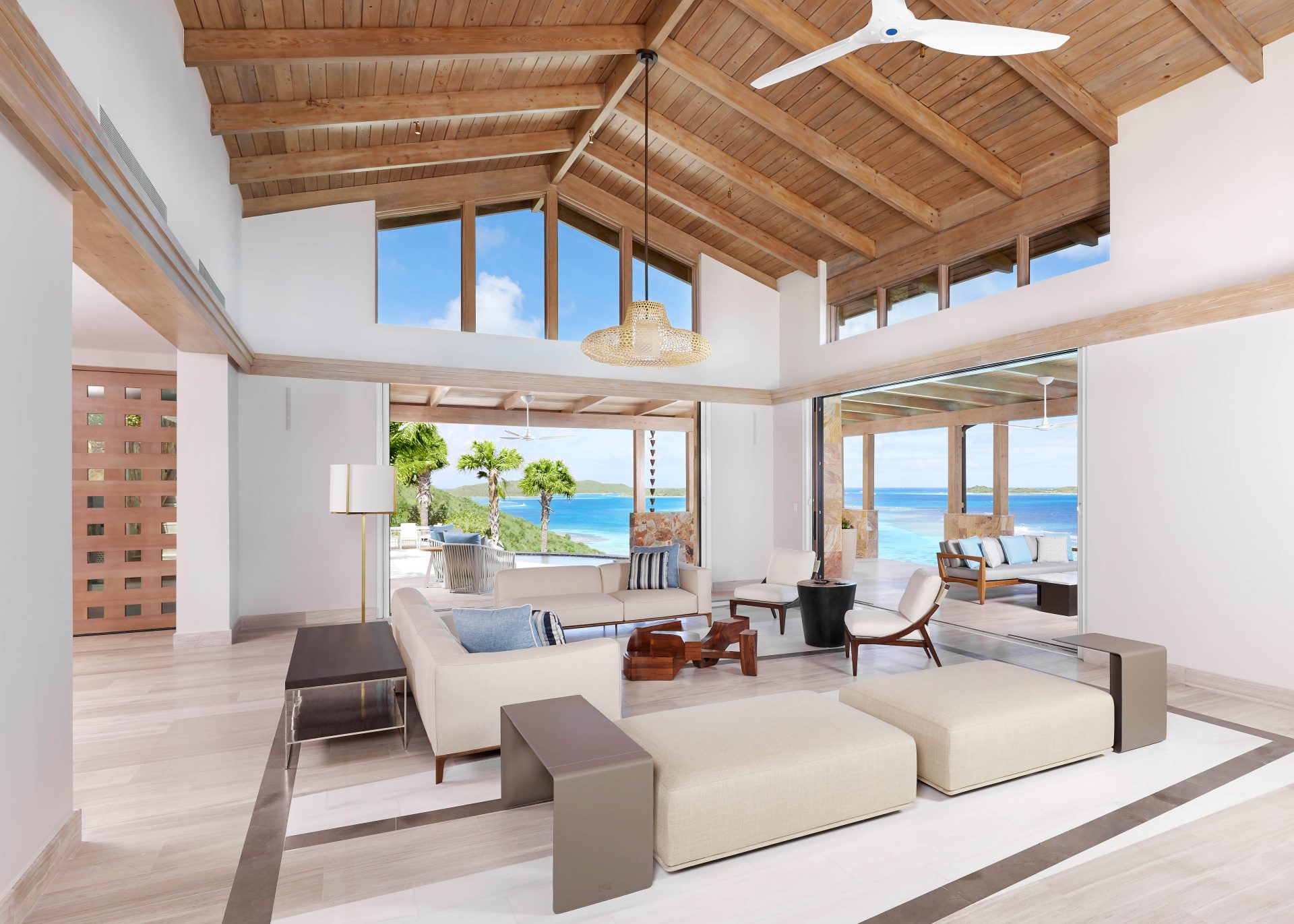 A modern living room in a Maronti villa features a high wooden ceiling and large windows offering a stunning ocean view. Cream-colored sofas, a wooden coffee table, and contemporary chairs furnish the space. A minimalist ceiling fan and pendant lights hang gracefully from the ceiling.