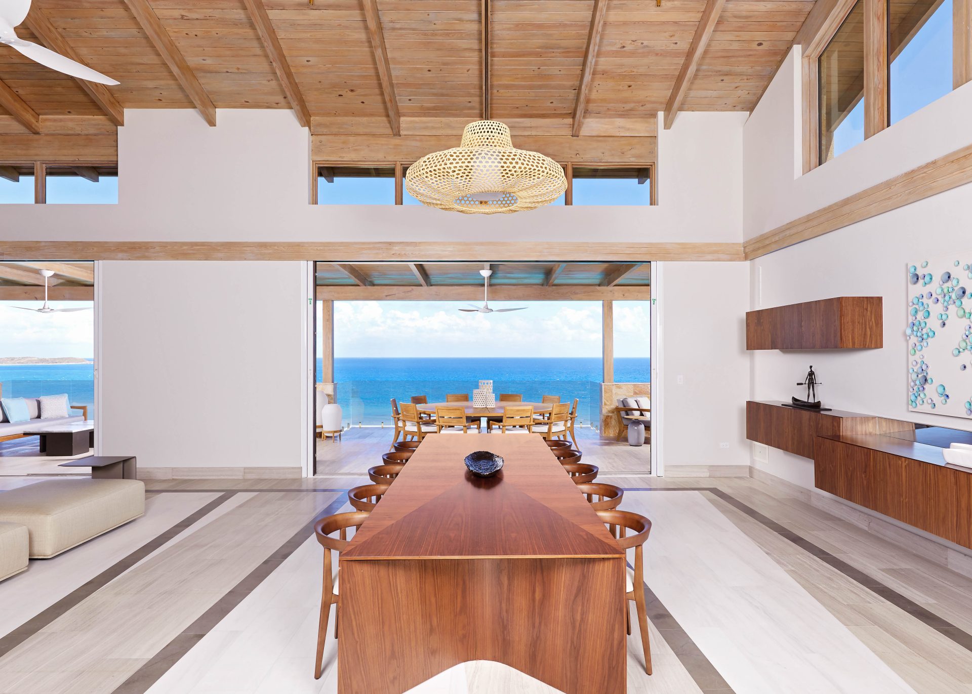 A spacious, open-plan room in the Maronti Villa with a long wooden dining table and chairs leading to a patio overlooking a bright blue ocean. The room features high wooden ceilings, modern minimalist decor, and a large woven chandelier. A lounge area is visible on the left.