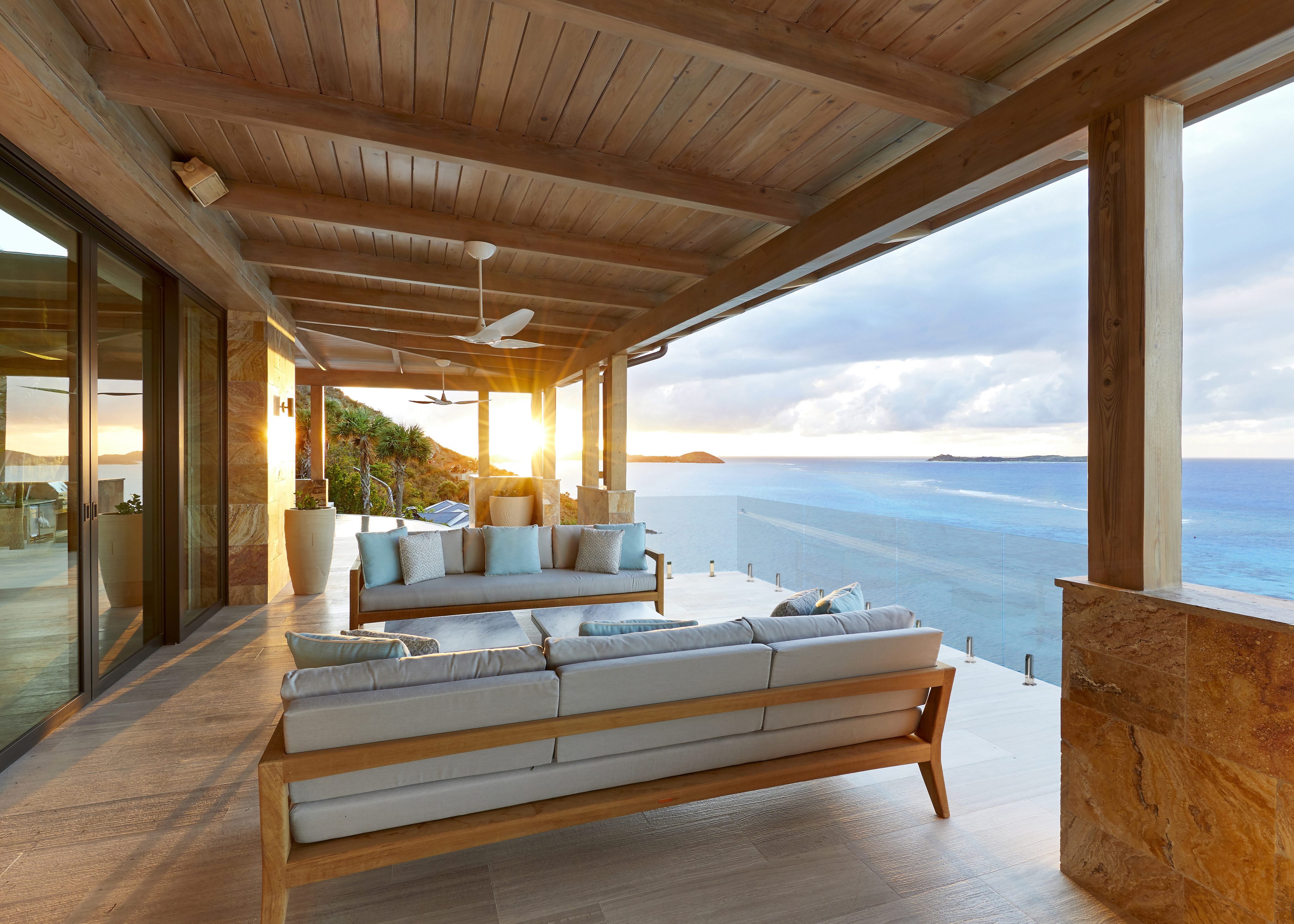 A luxurious outdoor patio at Maronti Villa overlooks a scenic ocean view at sunset. The space features wooden beams, light grey sofas with blue and white cushions, large floor-to-ceiling windows, a ceiling fan, and stone columns. The horizon blends sea and sky beautifully.