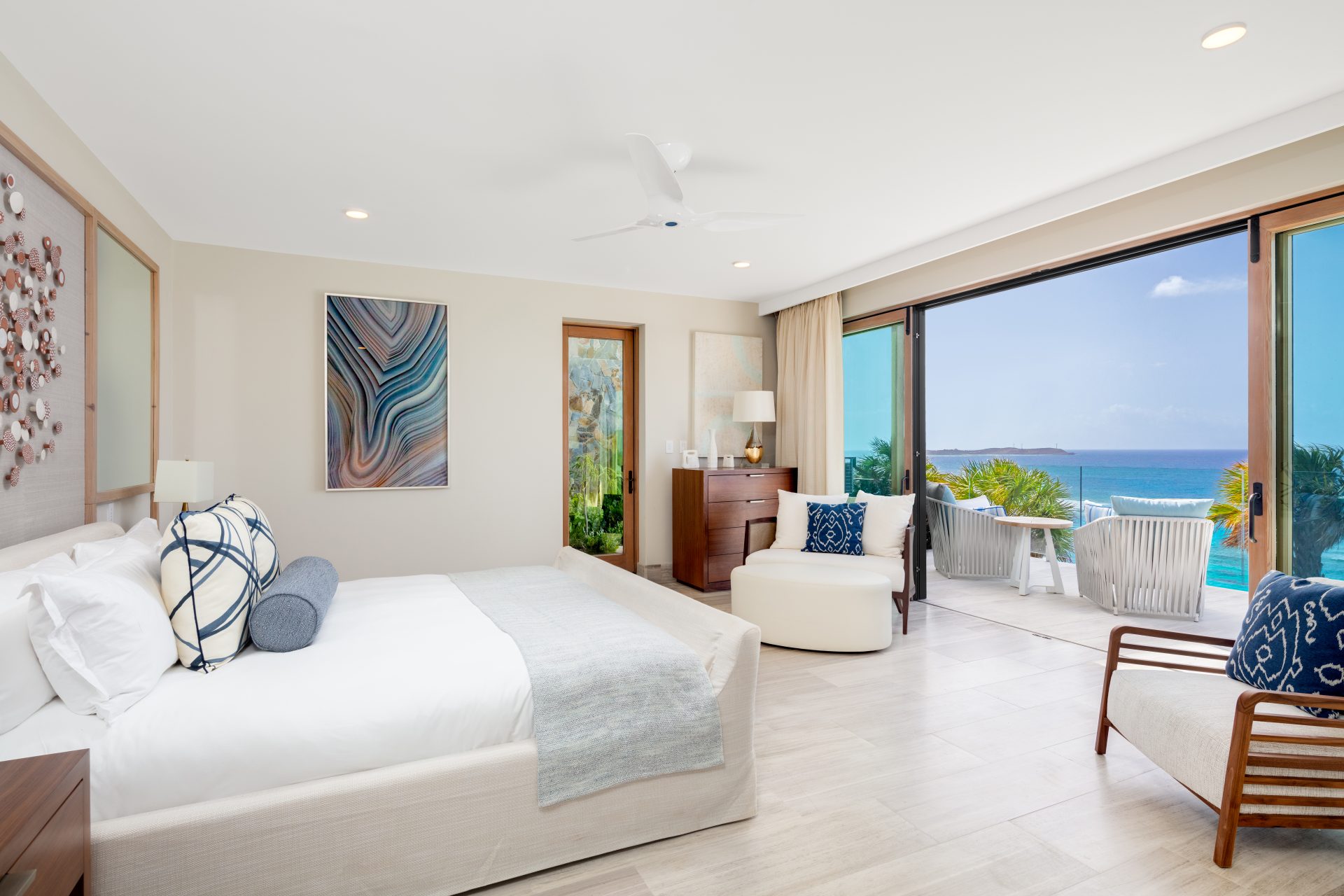 A spacious, modern bedroom in the Maronti villa with white walls and light wood flooring features a large bed with white linens and blue accents. The room has sliding glass doors opening to a patio with chairs overlooking an ocean view. Decor includes abstract art and minimalist furnishings.