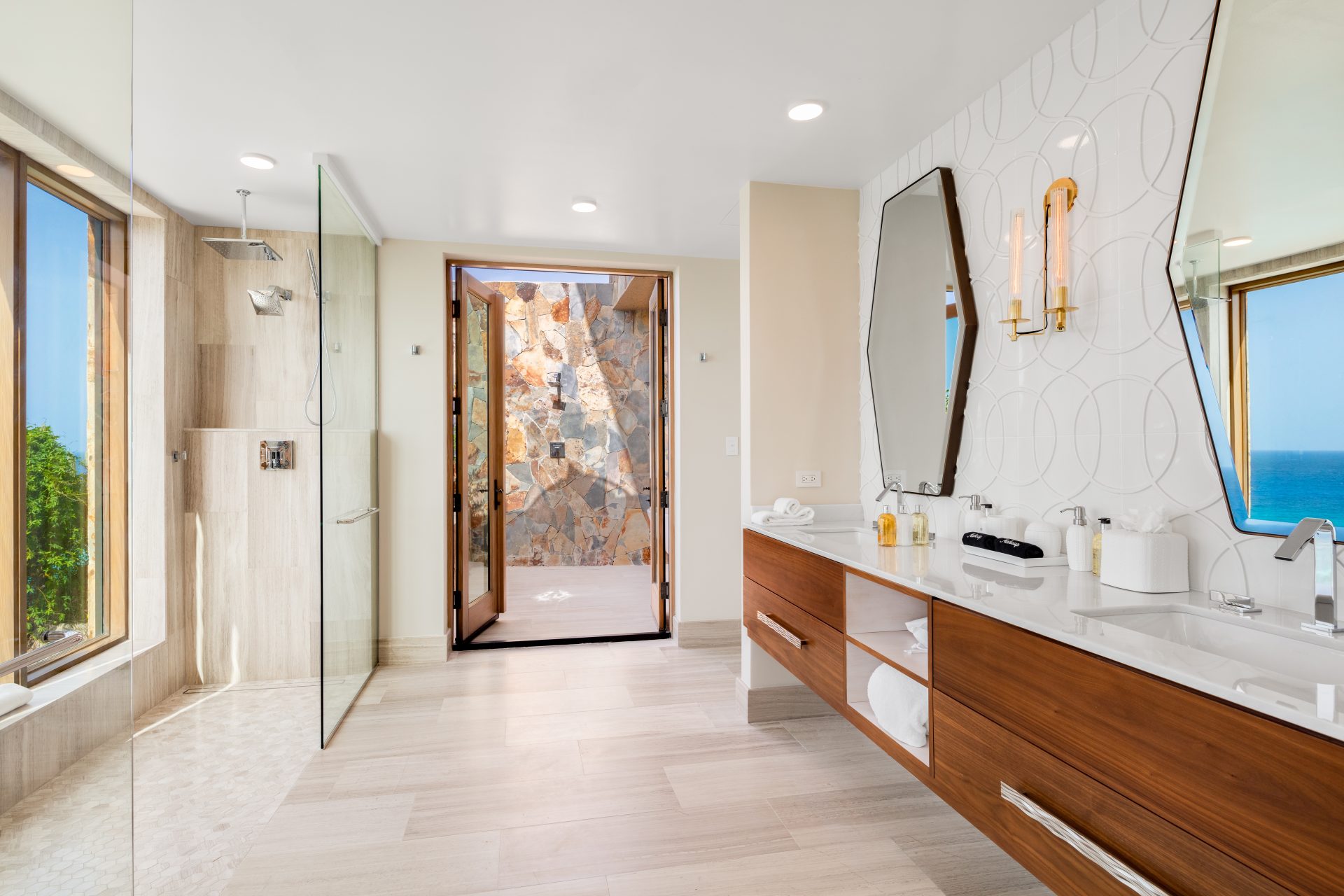 Modern bathroom in a Maronti villa with large windows showcasing ocean views. It features a walk-in shower with glass doors, dual vanity with hexagonal mirrors and contemporary light fixtures, wooden cabinets, and beige tiled flooring. An outdoor rock wall is visible through an open door.