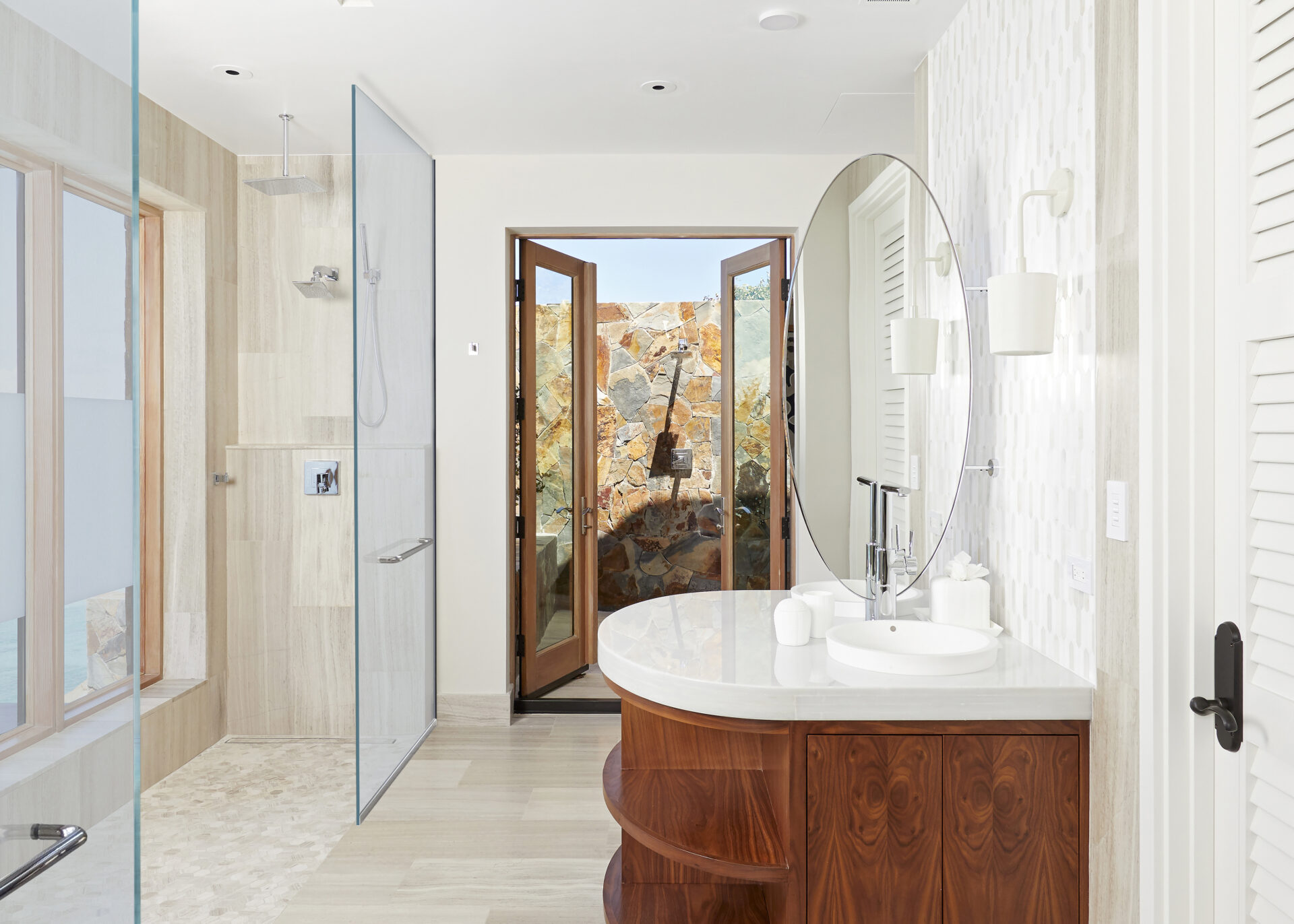 Modern bathroom in a luxurious villa featuring a glass shower, a circular mirror above a wooden vanity, and double doors leading to an outdoor area with stone walls reminiscent of Maronti. The space is bright with neutral tones and elegant fixtures.