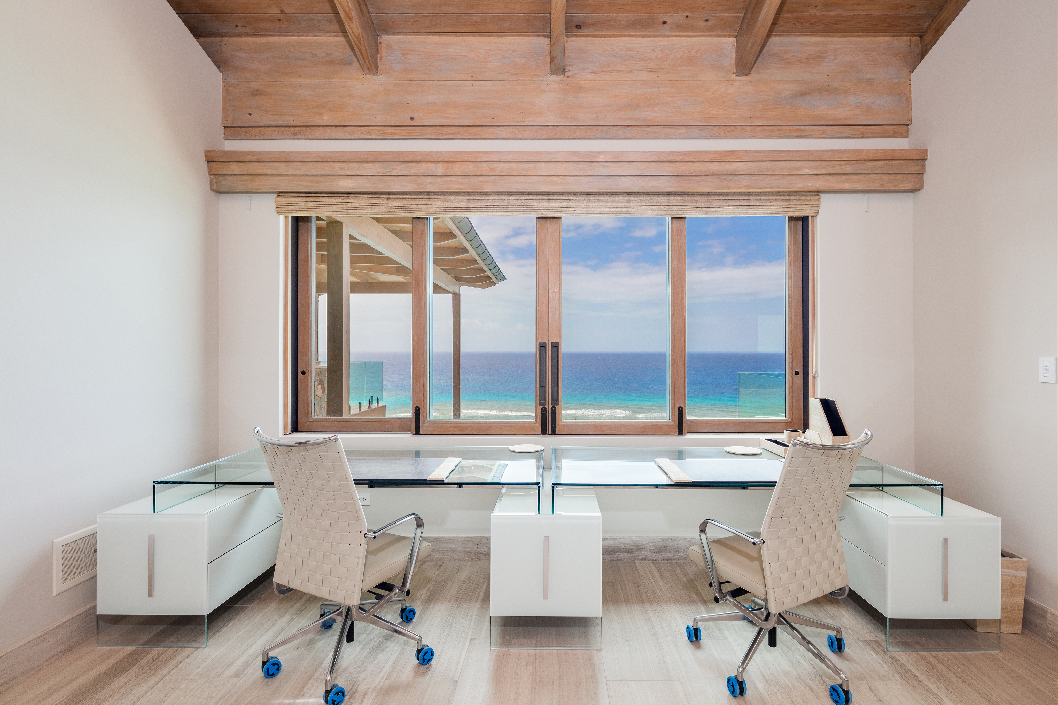 The modern office space in Villa Maronti features two glass desks and chairs facing a large window overlooking the ocean. With wood accents and a slanted ceiling, this serene room offers a spacious work environment perfect for creativity and focus.