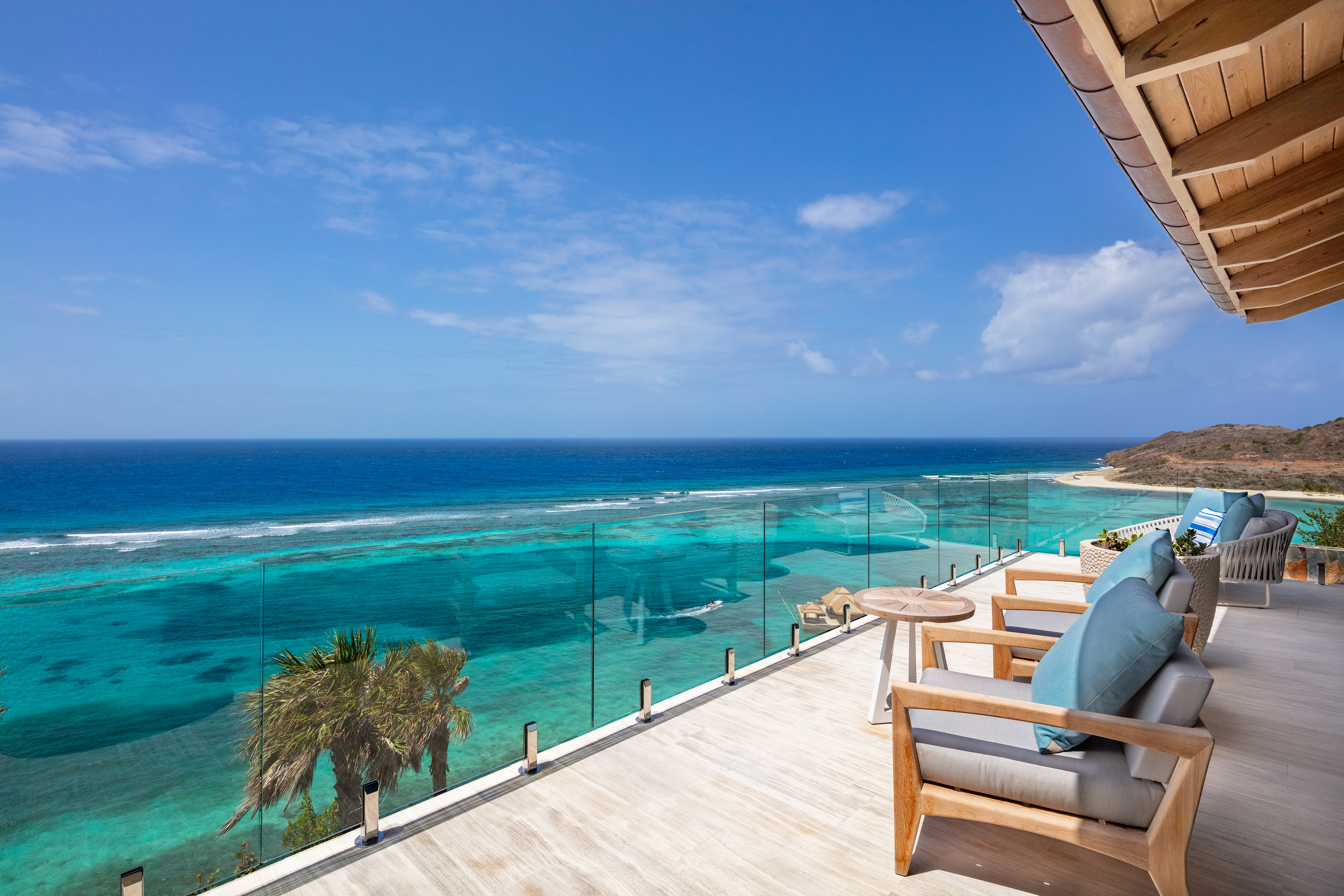 A spacious balcony at Villa Maronti offers a stunning ocean view with clear blue waters stretching to the horizon. Comfortable chairs with cushions are arranged on the wooden deck, framed by a glass railing, under a partly cloudy sky.