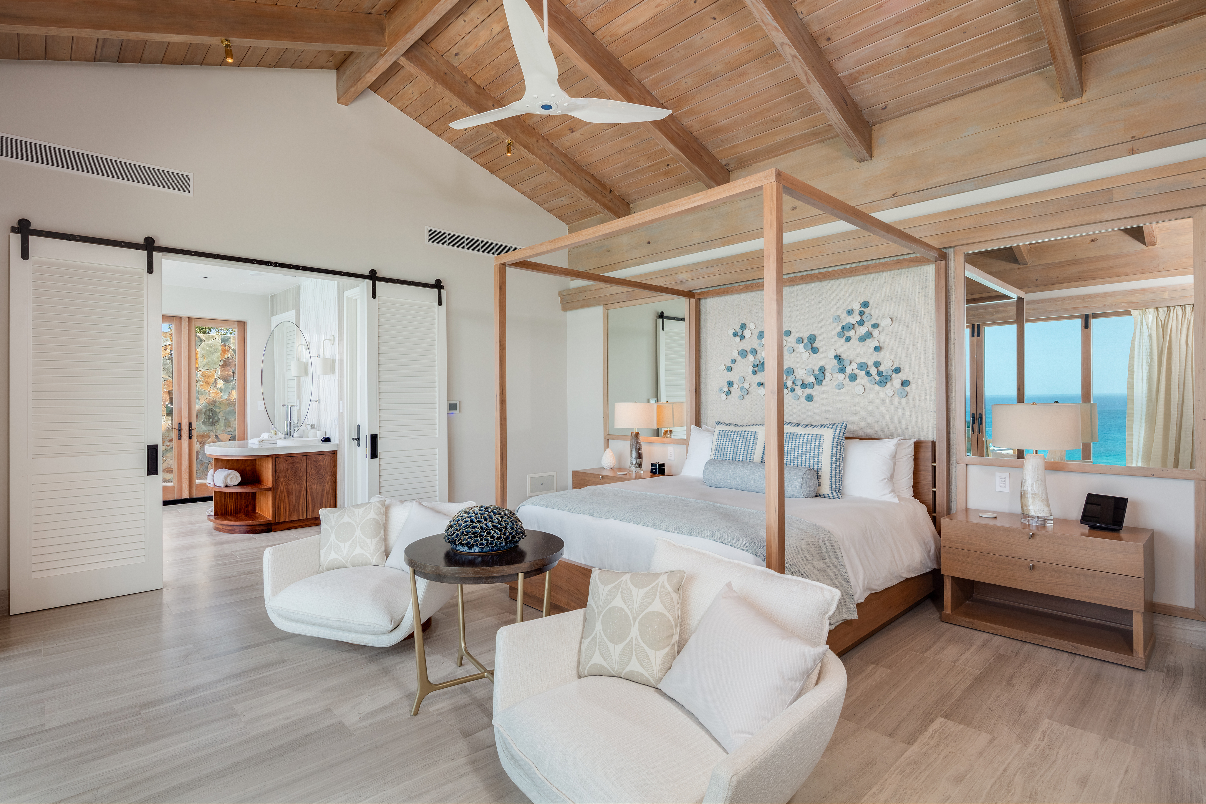 The luxurious Villa Maronti bedroom features a wooden ceiling and a large four-poster bed with white and blue bedding. Two white armchairs and a wooden table sit in the foreground. Open doors reveal a modern bathroom, complemented by an ocean view.