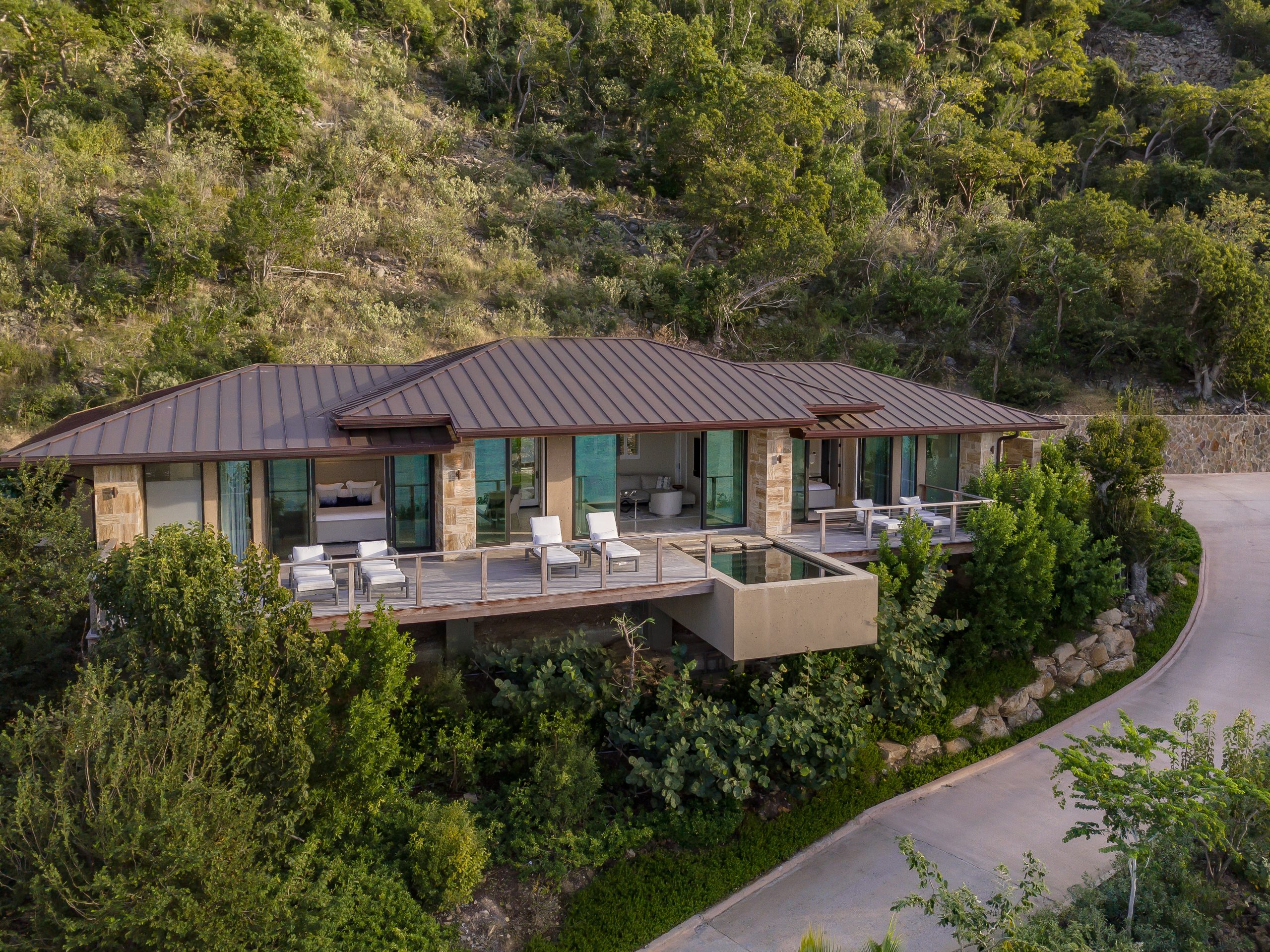 A modern villa with a metal roof sits on a hillside surrounded by lush green vegetation. The house features large windows, a spacious deck with outdoor furniture, and a small, elevated pool that extends from the deck. A driveway curves to the right, reminiscent of something MoneyPenny might admire.