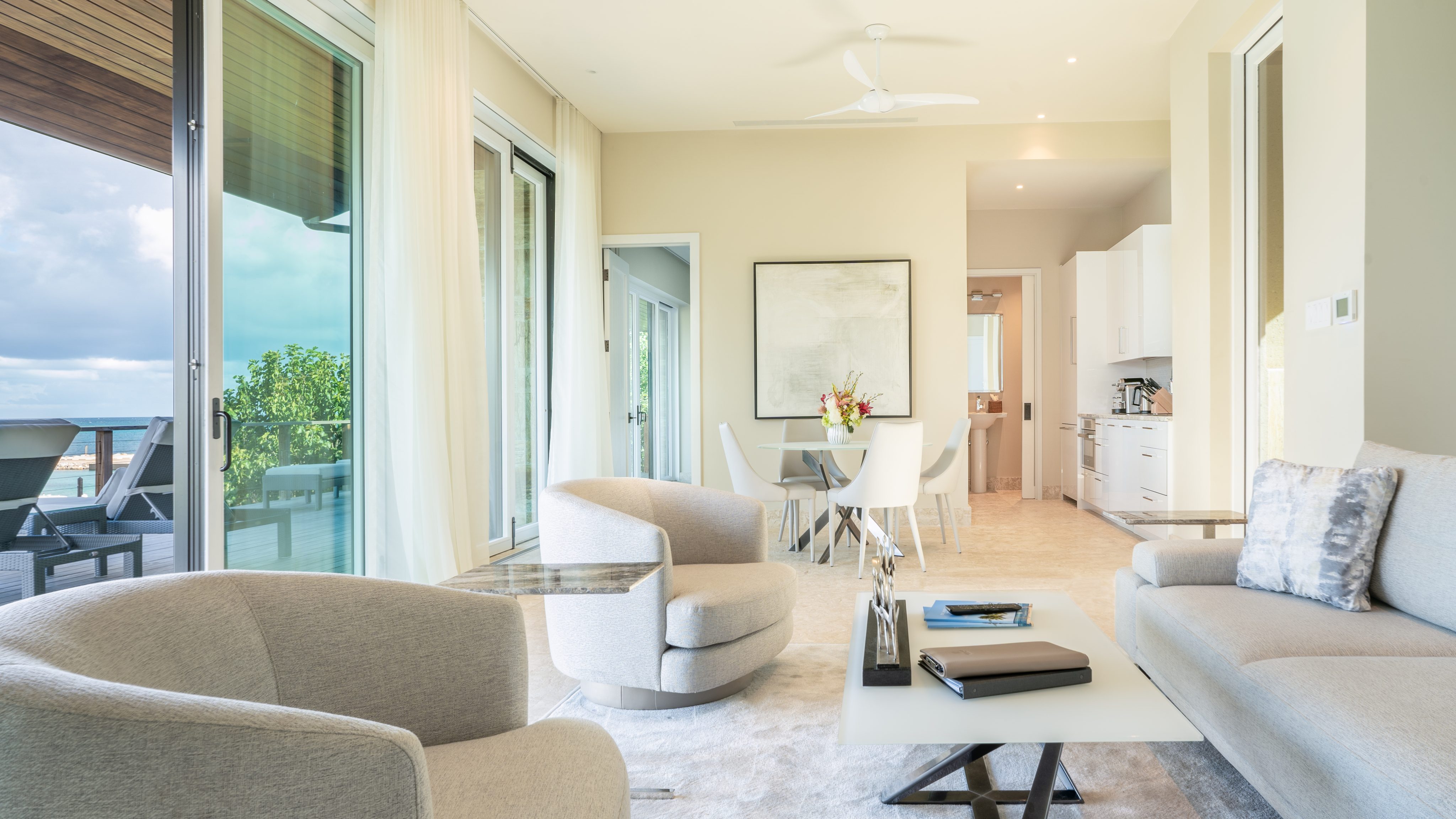 At MoneyPenny Villa, the modern living room boasts neutral tones with two armchairs and a sofa around a white coffee table. The background reveals a dining area with a table and chairs. Large windows draped in sheer curtains invite natural light, and a balcony view is visible on the left.