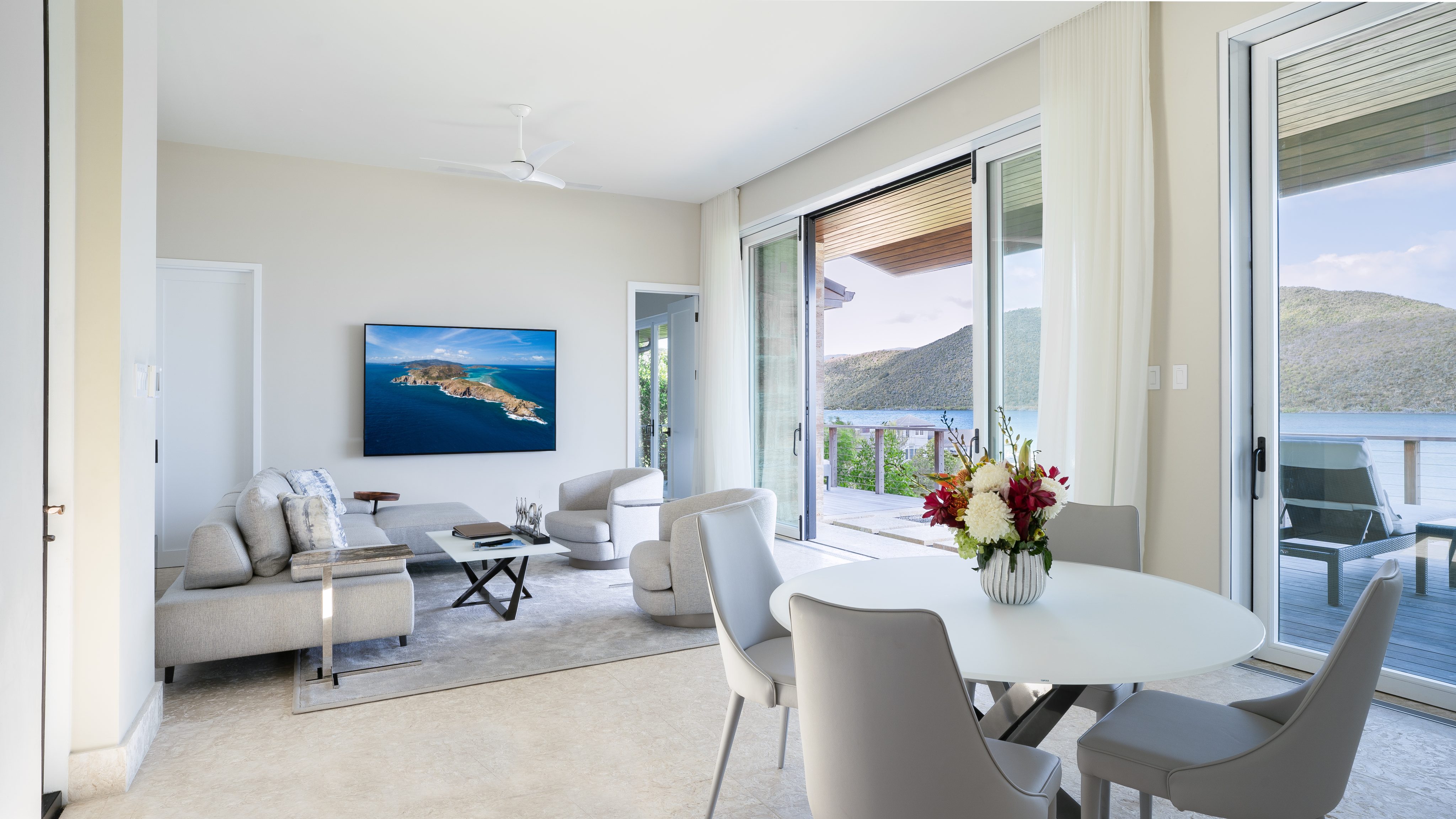 A modern living and dining room in a luxurious villa with light-colored walls and floor. The living area has a grey sofa, armchairs, a coffee table, and a wall-mounted TV. The dining space features a white round table with grey chairs and a vase of flowers. Large windows open to a balcony with scenic views of hills and water.