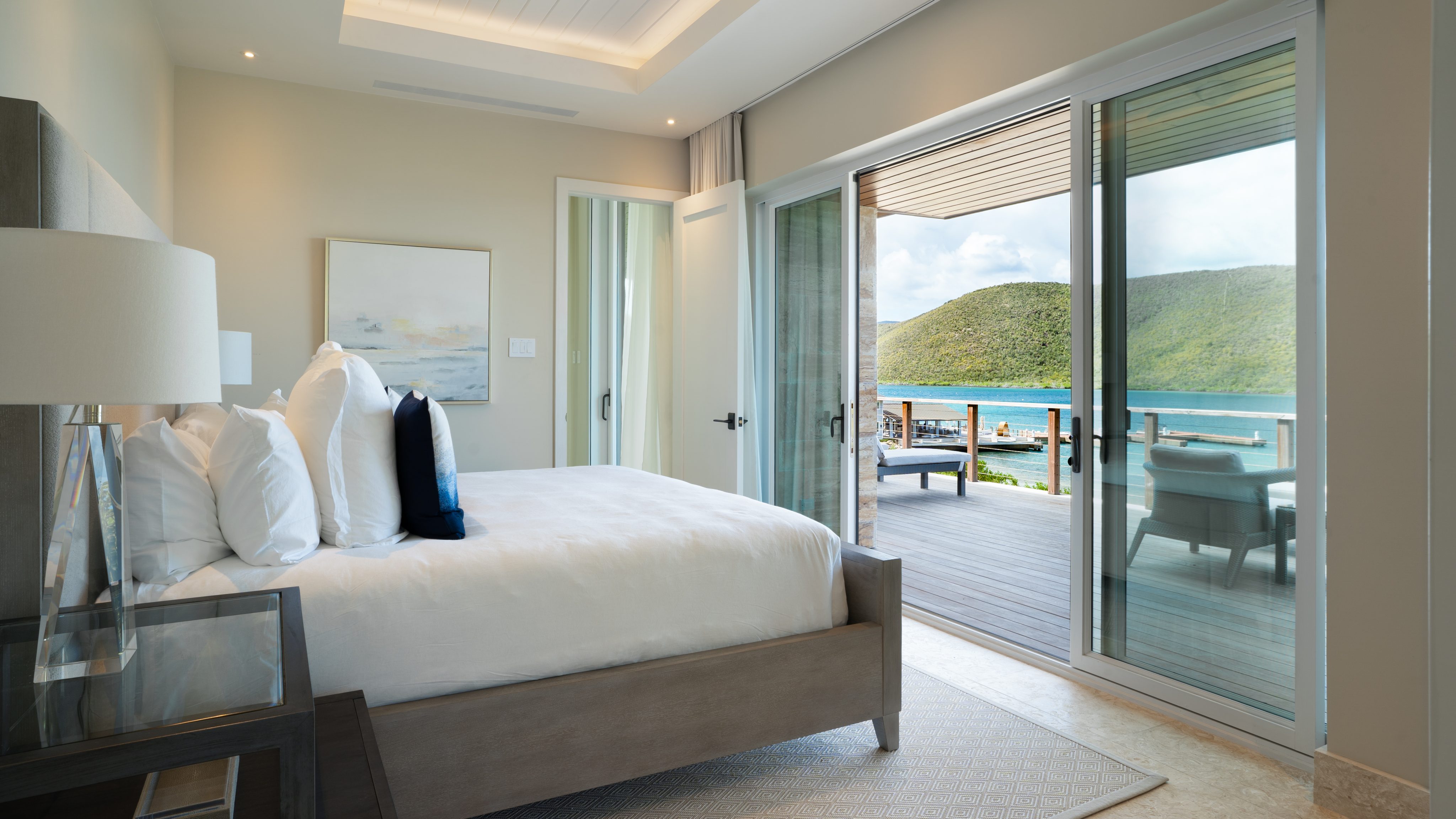 A modern, airy bedroom at MoneyPenny Villa features a large bed adorned with white linens and navy blue accent pillows. Glass sliding doors open to a balcony with a scenic view of a serene lake and lush green hills, blending luxury accommodation with neutral decor and ample natural light.