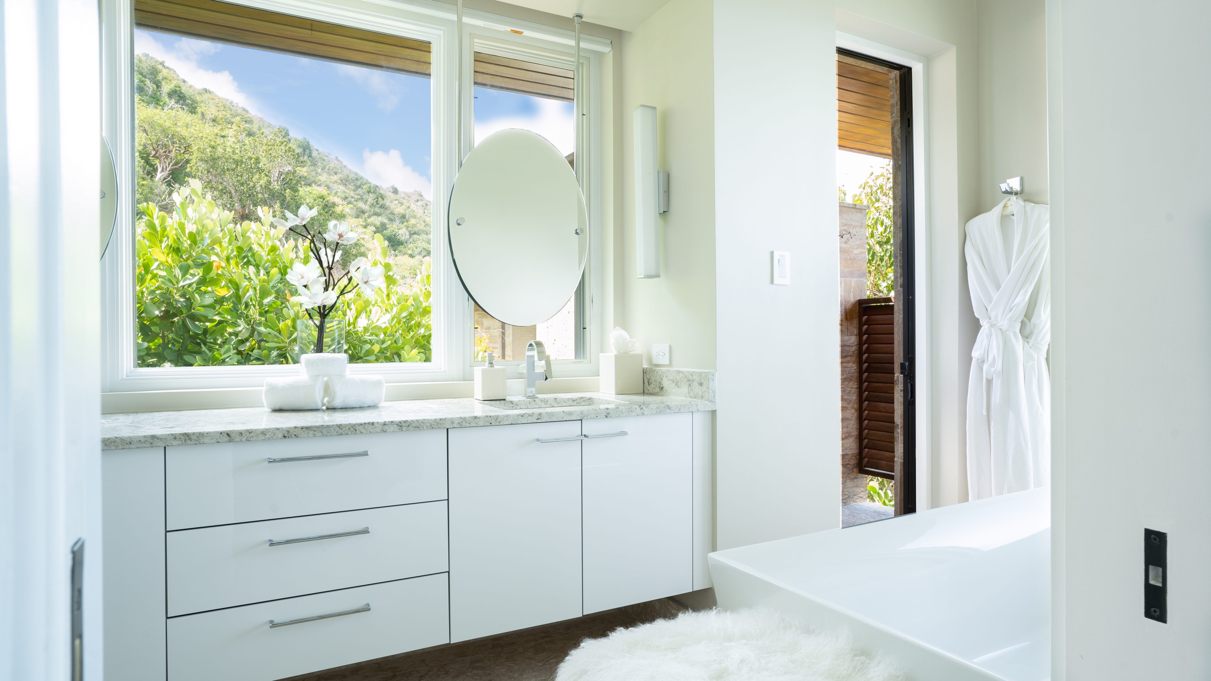 A modern bathroom in MoneyPenny Villa boasts a large window overlooking lush greenery. The room features a white countertop with two sinks, a round mirror, a white bathrobe hanging near the open door, and a fluffy white rug. Natural light fills the space, creating a bright and airy feel.