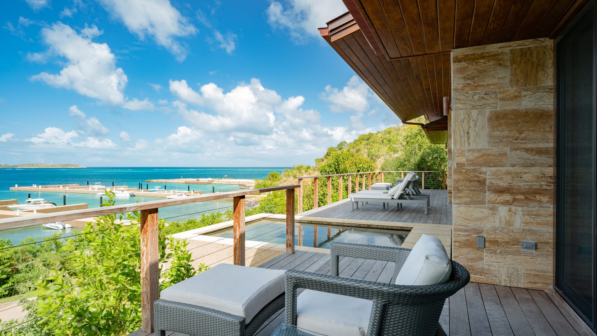 A seaside balcony at the luxurious villa features cushioned lounge chairs overlooking a tranquil marina. The view includes blue skies, scattered white clouds, green hills, and the crystal-clear ocean. A stone wall and wooden railing frame this modern, inviting space reminiscent of MoneyPenny's refined aesthetic.