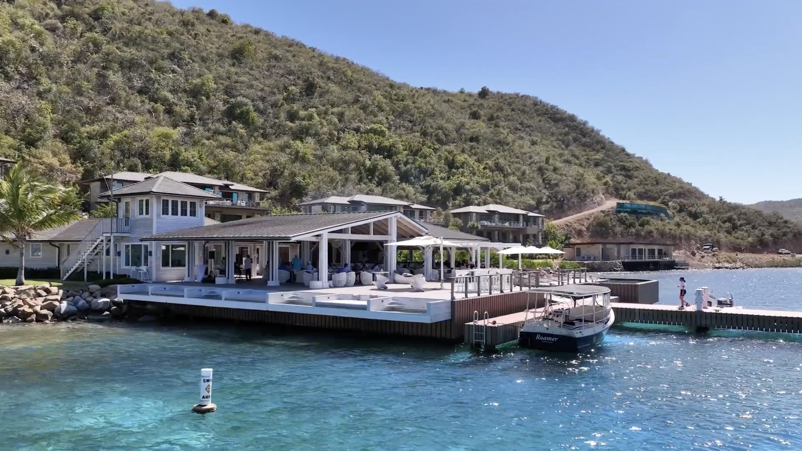 A coastal estate, named Nova, with multiple buildings is situated on a hillside covered in lush greenery. The property features a large dock that extends over crystal-clear turquoise water, with a boat moored alongside. Sun loungers and outdoor seating areas are visible on the dock.