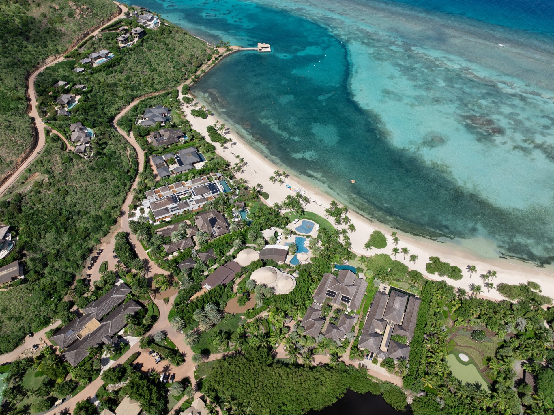 Aerial view of a tropical coastal resort with beach villas, a white sandy beach, several pools, and lush greenery. The clear blue ocean with coral reefs and a small docking pier are visible. Pathways wind through the resort, connecting various facilities and accommodations.