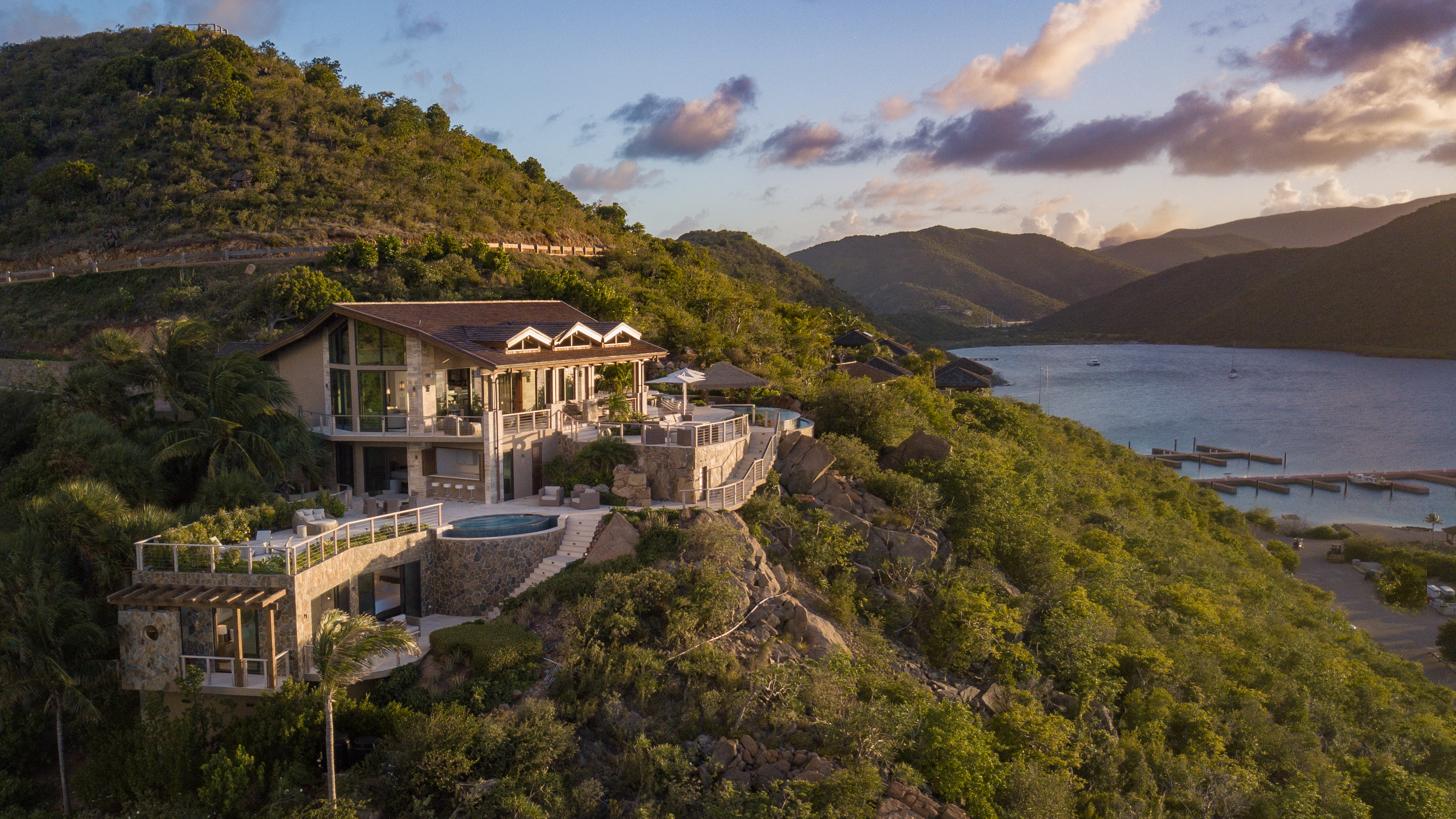 A luxurious hillside villa overlooking a tranquil bay, this real estate gem features a lush garden, a spacious pool deck, and expansive terraces with breathtaking ocean and mountain views. The sun sets, casting a warm golden glow over the verdant landscape—a true dream property.