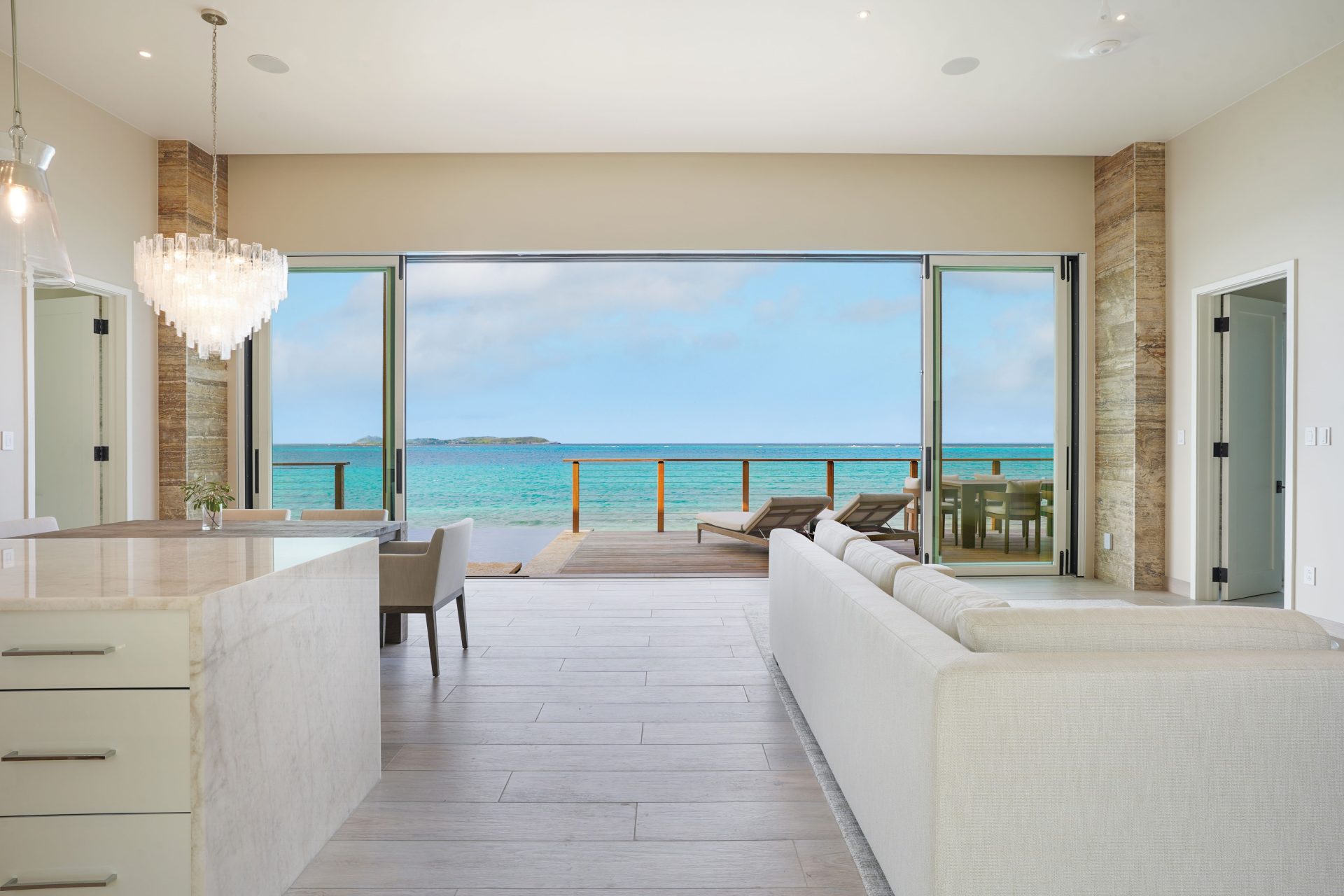 A modern, open-concept living space with light-colored furniture and a kitchen island leads to a wide, open sliding door. Outside, there's a wooden deck with lounge chairs and a dining table, offering a stunning view of the turquoise ocean and clear blue sky.