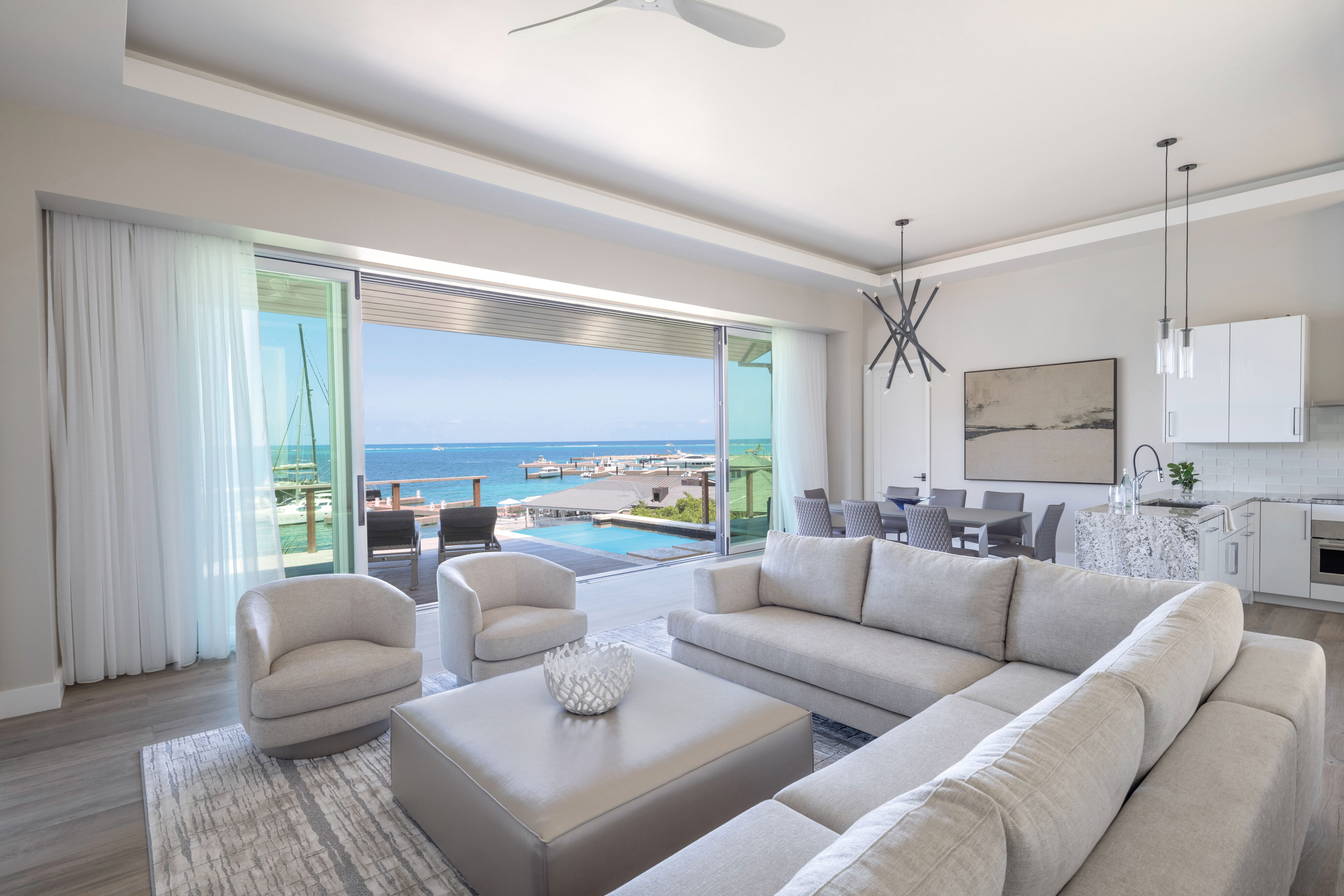 A modern living room with light gray furniture, including a large sectional sofa, armchairs, and a central ottoman. Floor-to-ceiling windows open to an outdoor patio with a pool and a breathtaking marina view beyond the clear blue ocean. The kitchen and dining area are visible on the right.