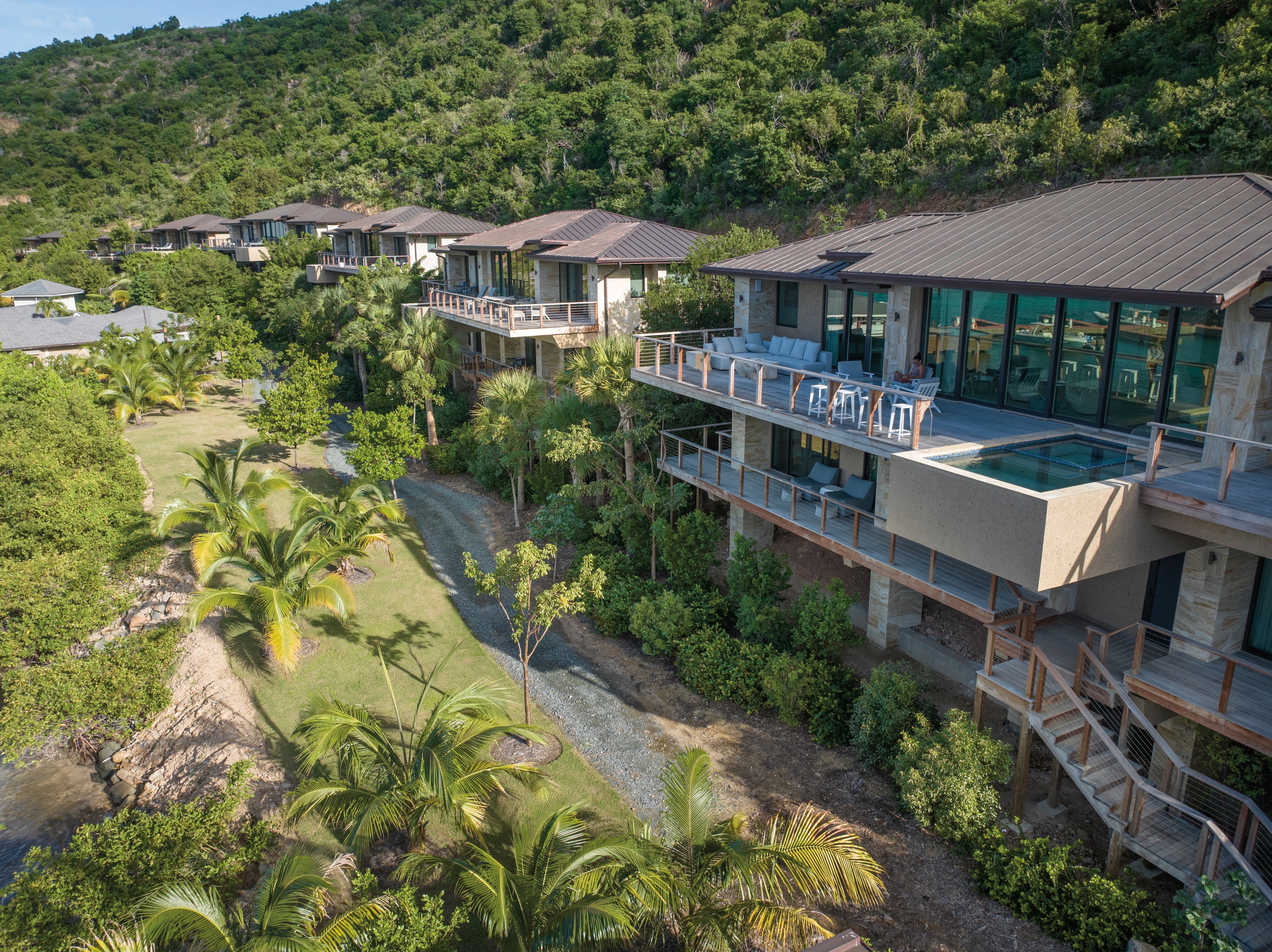 A series of modern hillside homes with large glass windows and balconies overlook a lush landscape with palm trees, leading to a grassy area by the marina. The houses blend into the scenic tropical surroundings, combining natural elements with contemporary architecture.